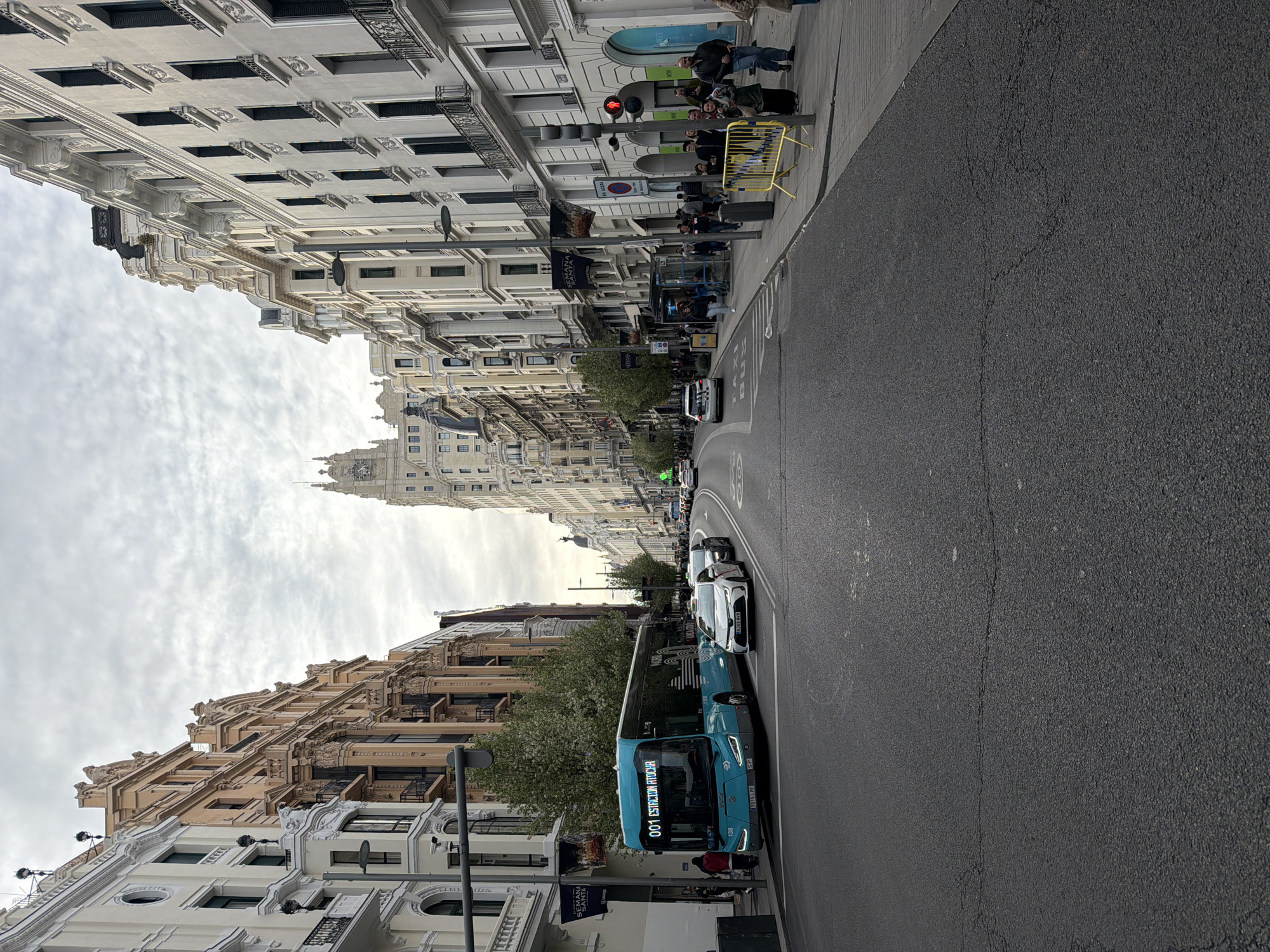The image depicts a street scene in Madrid, Spain. The camera faces down a straight avenue, lined on both sides with buildings of varying architectural styles, primarily featuring ornate, classical facades. The street is paved with dark asphalt, showing signs of wear. On the right sidewalk, a small crowd of people is waiting near a traffic light, which is red. There's a yellow barricade nearby. A bus is visible on the left side of the road, partially obstructing the view of some buildings. Several cars are positioned behind the bus, and others can be seen further down the avenue. The sky is overcast. At the end of the avenue, a tall, ornate building is visible, suggesting that the photo is likely taken along Gran Vía, known for its varied architectural styles.