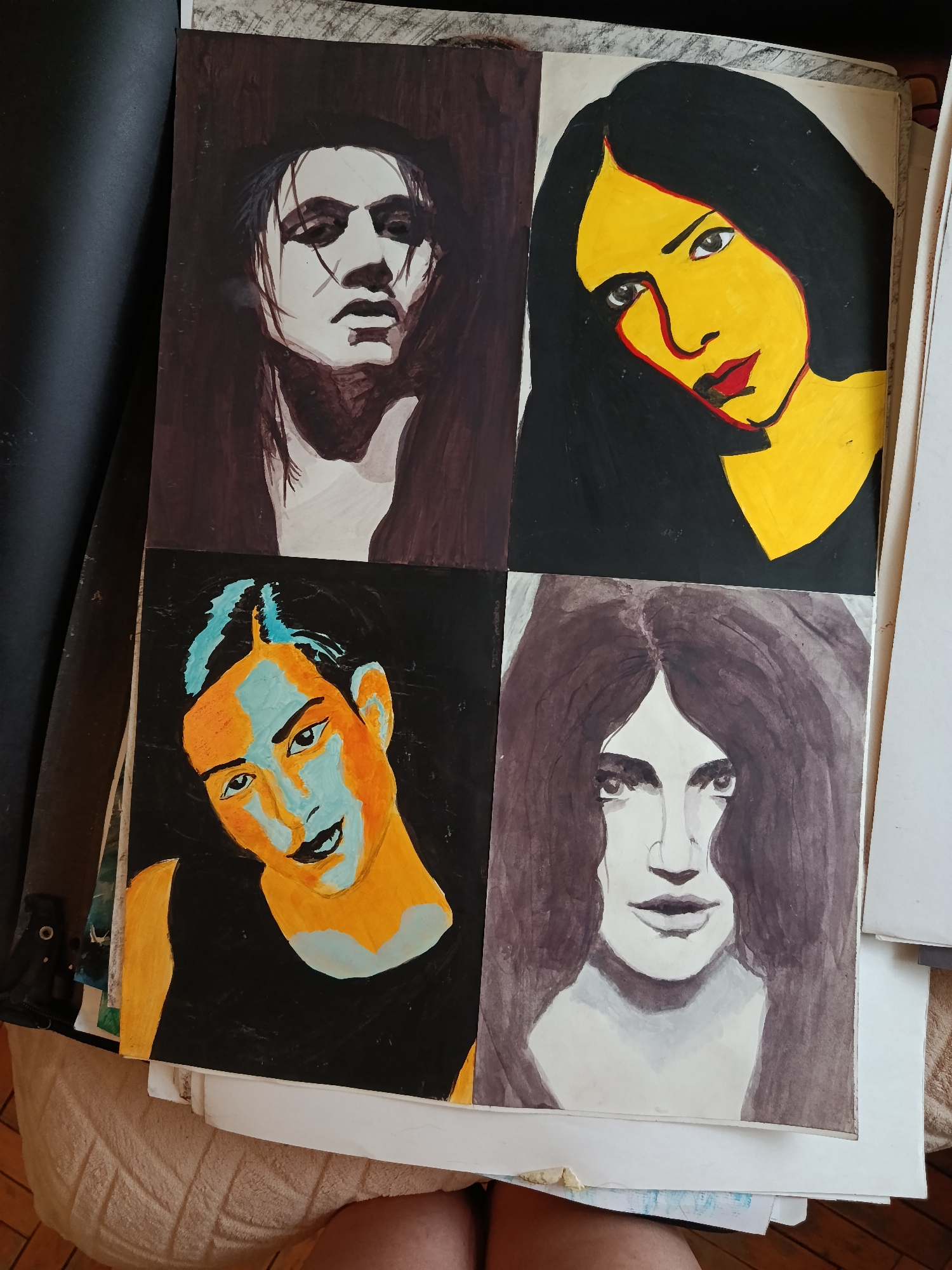 The image shows a piece of artwork with four portraits arranged in a square. The top left portrait is a black and white/brown monochromatic depiction of a person with long dark hair. The top right portrait is a woman with black hair, yellow skin, and red lips. The bottom left portrait is a woman with black hair, orange skin, and blue highlights. The bottom right portrait is a person with long dark hair, mostly monochromatic with some brown hues. The artwork appears to be resting on a stack of papers, next to a black object (possibly a notebook or portfolio) and a beige surface that could be a piece of furniture. There is no indication of location being in Moscow, Russia or any other specific place.