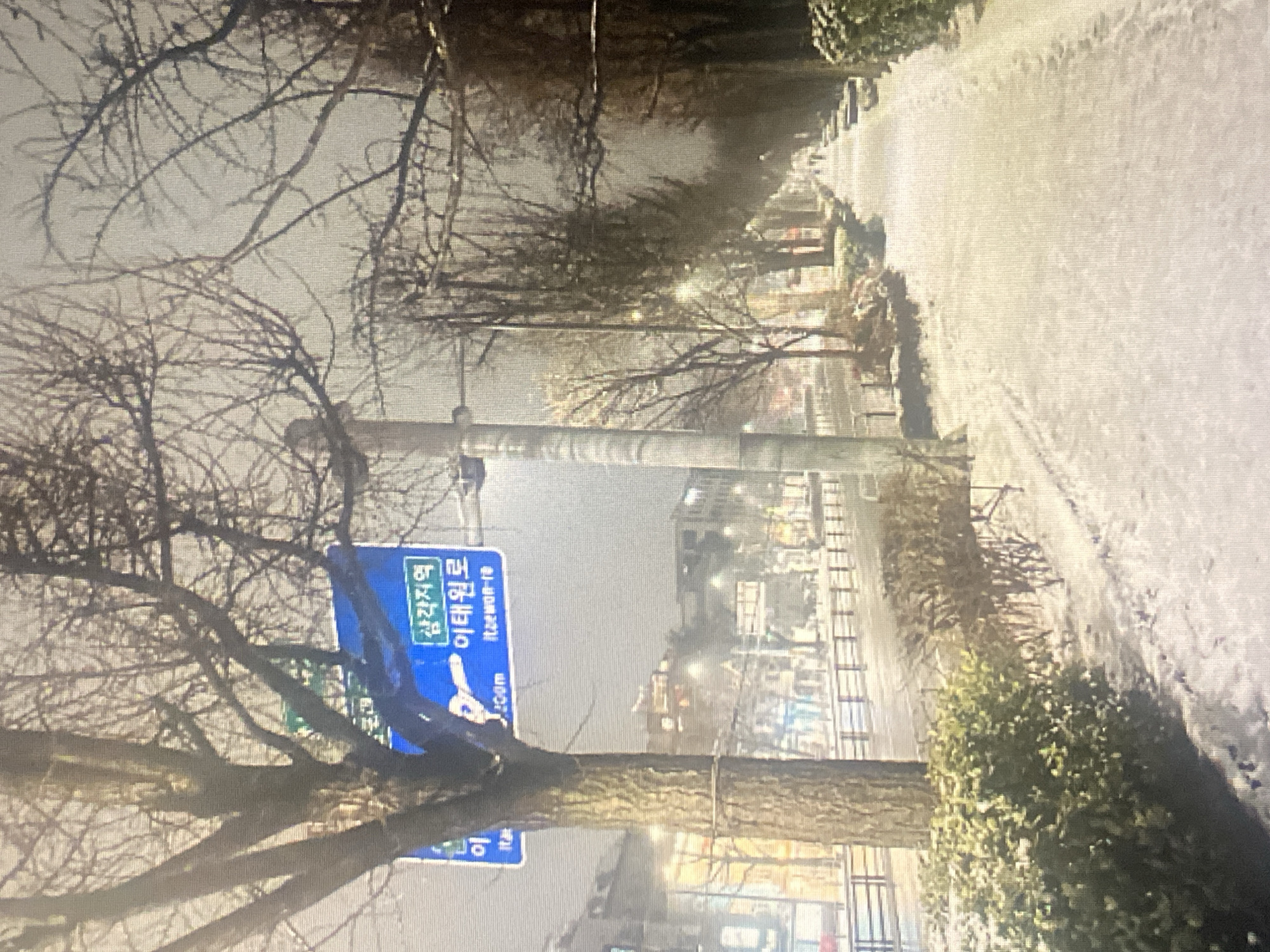 The image depicts a snow-covered street scene in an urban environment at night. A blue street sign with Korean text is visible, indicating directions to "Samgakji Station" and "Itaewon-ro". Bare trees line the sidewalk, which is covered in snow. A road runs alongside the sidewalk, with a railing and some buildings in the background. The overall impression is a cold, winter evening in a city.