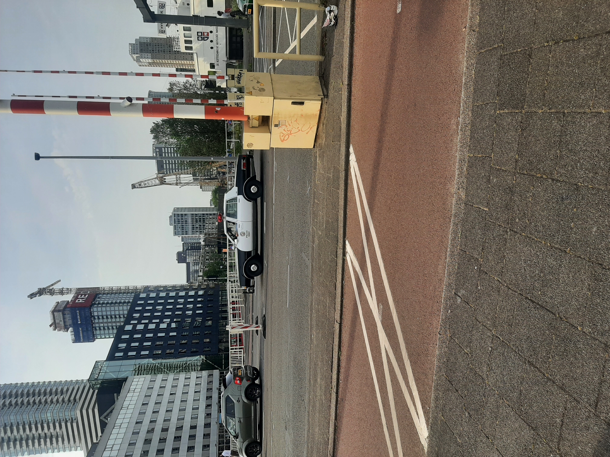 The image shows a street scene in Rotterdam, Netherlands, likely near a port or industrial area. There are several modern high-rise buildings under construction, including one with a crane on top. A white service vehicle with a black bumper is parked along the road, near a closed railroad crossing with red and white striped barriers. A small green car is parked further down the street. There are light-colored wooden crates on the sidewalk with graffiti. The street has a gray asphalt surface, and the sidewalk has a reddish-brown bike lane separated by white lines.