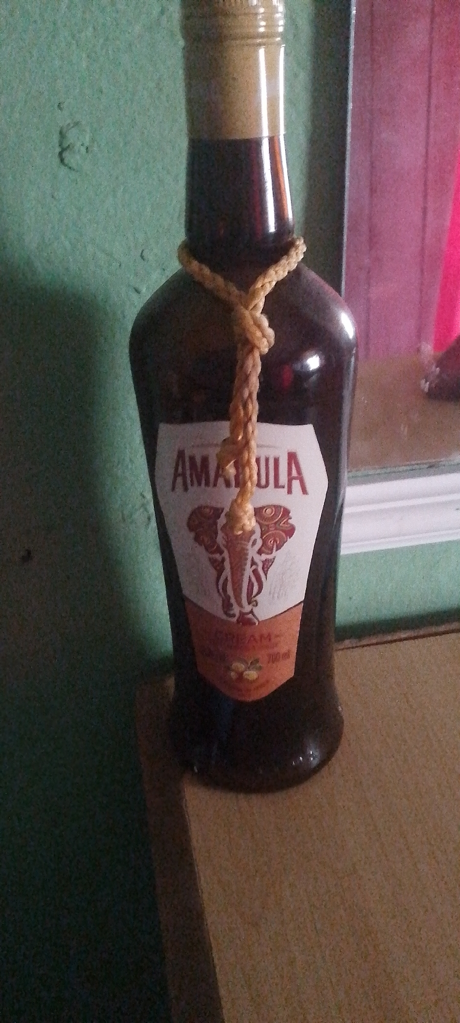 A bottle of Amarula Cream liqueur stands on a wooden surface. The bottle is dark brown glass with a light-colored neck and cap. A braided tan rope is tied around the neck of the bottle. A white label on the bottle features a red outline and an image of an elephant. The words "AMARULA" and "CREAM" are visible on the label, along with "700 ml". The background consists of a textured green wall to the left and a blurred pinkish and white surface to the right, possibly a mirror or reflective material. The lighting is ambient, casting some reflections on the bottle. There are no people or animals in the image.