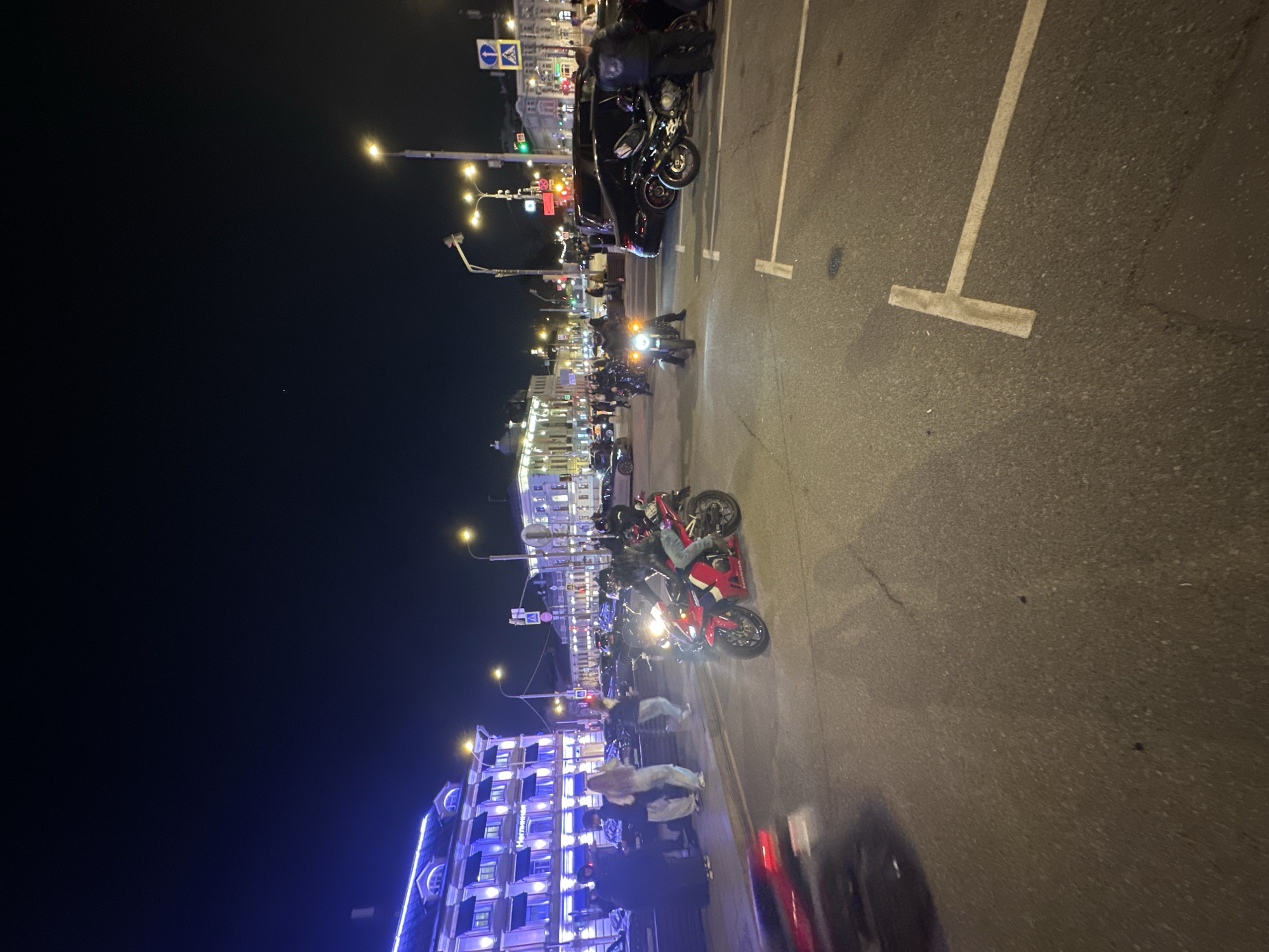 Here is a description of the image:
The image shows a nighttime scene in Moscow, Russia, with motorcycles parked along a street in the city center. The scene appears to be bustling with activity, as there are numerous people and motorcycles visible in the frame. The buildings are well-lit with street lights and other commercial lightings, especially illuminating the city center. The street has parking lane markings. Several tall buildings are in the background. Pedestrians are present.