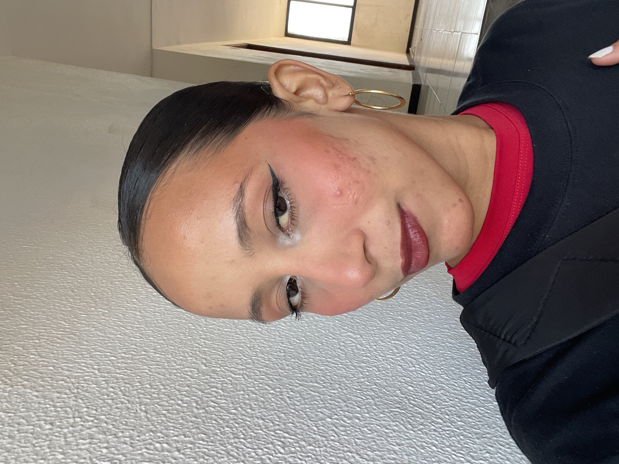 A medium shot captures a woman in a self-portrait, leaning her head against a textured, off-white wall. She is wearing a black crewneck sweatshirt over a red garment, with a black strap visible across her shoulder. The woman's dark hair is pulled back tightly. Her makeup includes a bold black winged eyeliner, light eyeshadow, noticeable blush on her cheeks, and dark berry lipstick. She wears a large, gold hoop earring on her right ear. The background includes a hallway with a tiled floor, a doorway, and a window. The woman's facial features include prominent cheekbones and visible acne scarring on her left cheek. The lighting appears to be natural, casting soft shadows. The scene suggests an indoor environment, possibly a residential hallway. This location is identified as being in Porlamar, Venezuela.