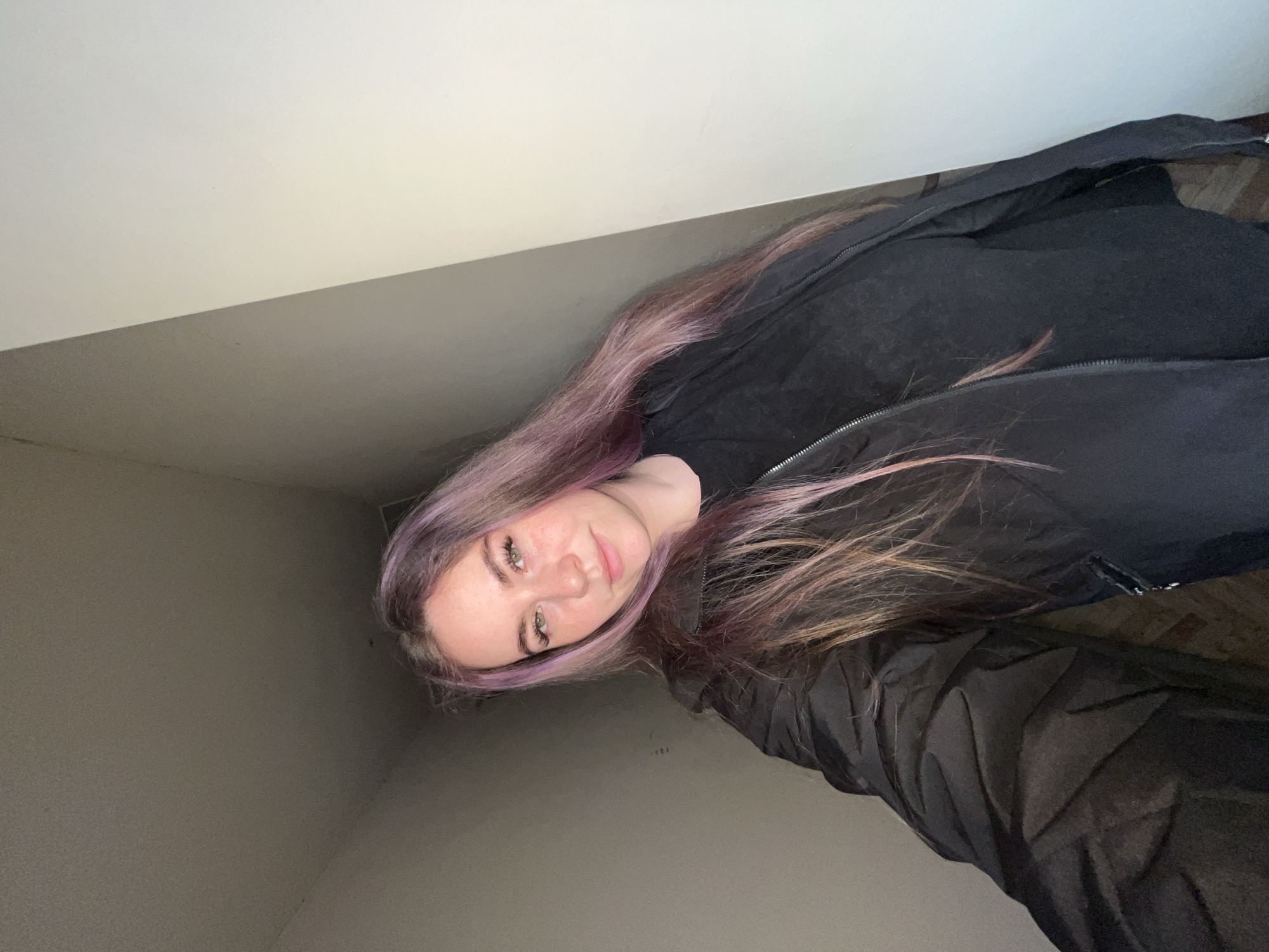 The photograph shows a young woman with shoulder-length, light brown hair with purple dye at the roots. She has fair skin, light eyes, and is looking directly at the camera with a neutral expression. She's wearing a black t-shirt and a black jacket with a visible zipper. The background appears to be an indoor space with light grey walls and a white ceiling, suggesting a room. It's not possible to determine the location in Chaikovsky, Russia based on the visual information in the image.