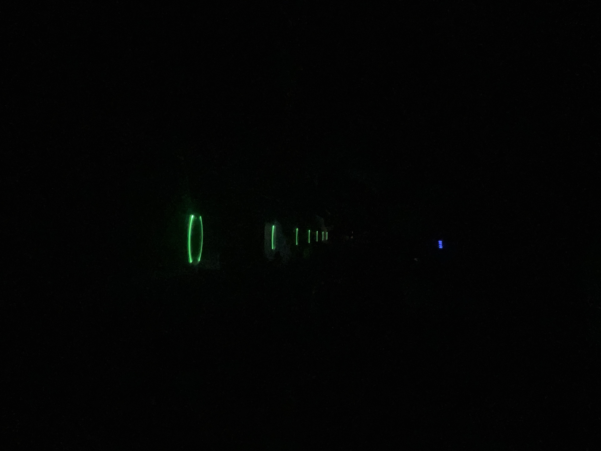 The image shows a series of lights, mostly green, diminishing in brightness and size with distance. The first light is a distinct green loop, followed by several green vertical bars. Further away, there's a small, faint blue light. The scene is extremely dark. No people or other identifiable objects are visible besides the lights. There is no information visible that identifies this location as Vinogradets, Bulgaria.