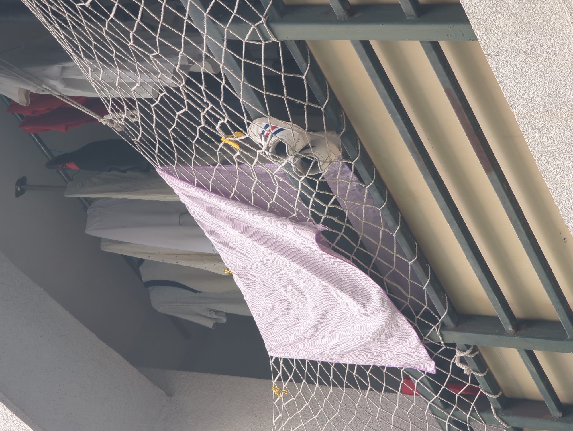 A safety net secured to a metal frame protects objects, including clothing, and a shoe from falling from an apartment in Shunde District, China. A lavender towel is clipped to the net, alongside a white shoe with red and blue stripes. The clothing appears to be hanging on a rod. The environment suggests an urban setting, with structural elements of the building visible.