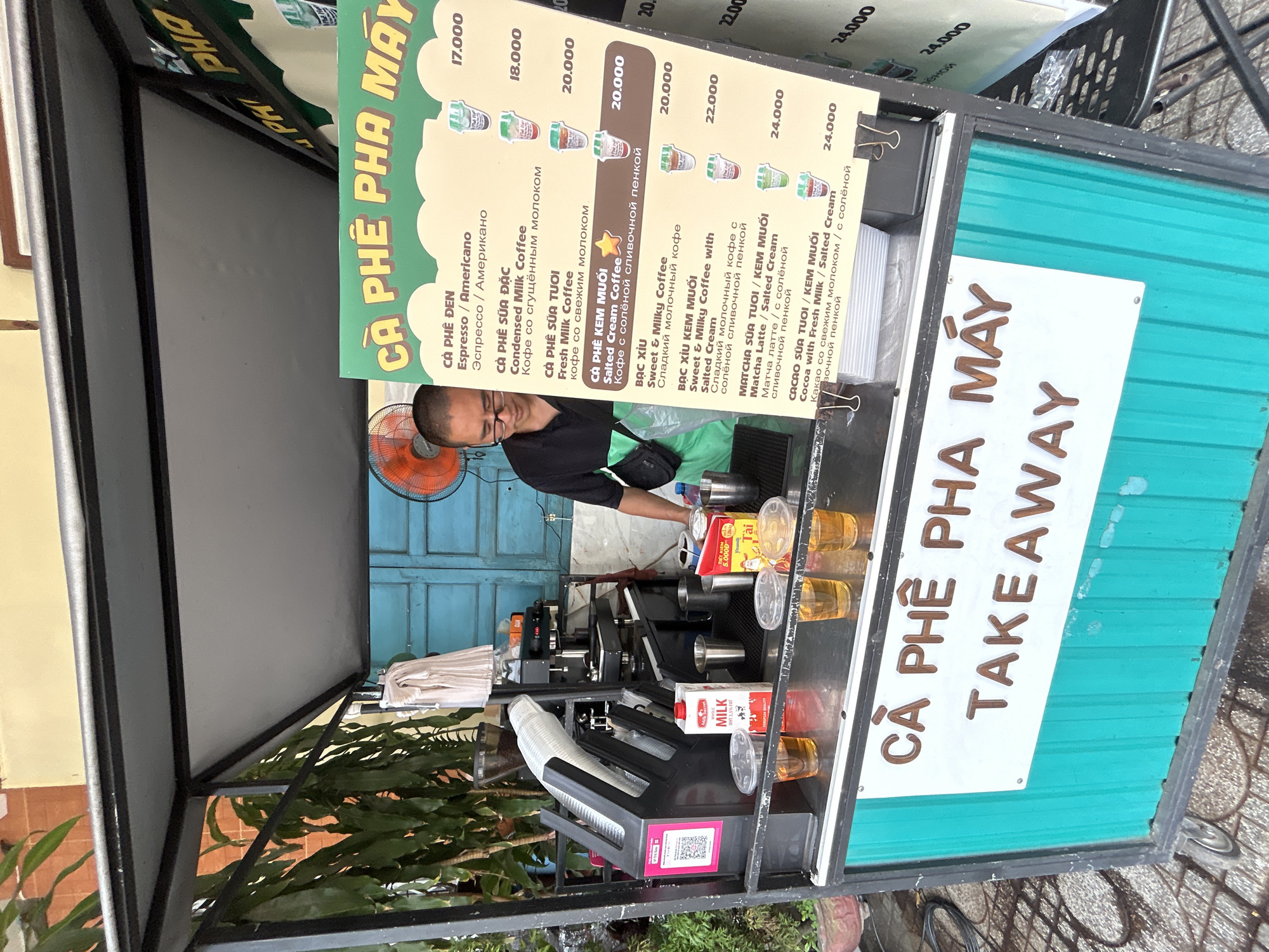 The image shows a coffee stand in Bac Natural Heritage Area Trang, Vietnam. A man is preparing drinks behind the counter. The stand has a menu displaying various coffee drinks with prices, including Espresso, Condensed Milk Coffee, Fresh Milk Coffee, Salted Cream Coffee, Sweet & Milky Coffee, and Matcha Latte. The menu is written in Vietnamese and Russian. The sign on the stand reads "CÀ PHÊ PHA MÁY TAKEAWAY". There are cups, milk, and other drink-making equipment on the counter. The stand has a blue metal frame.