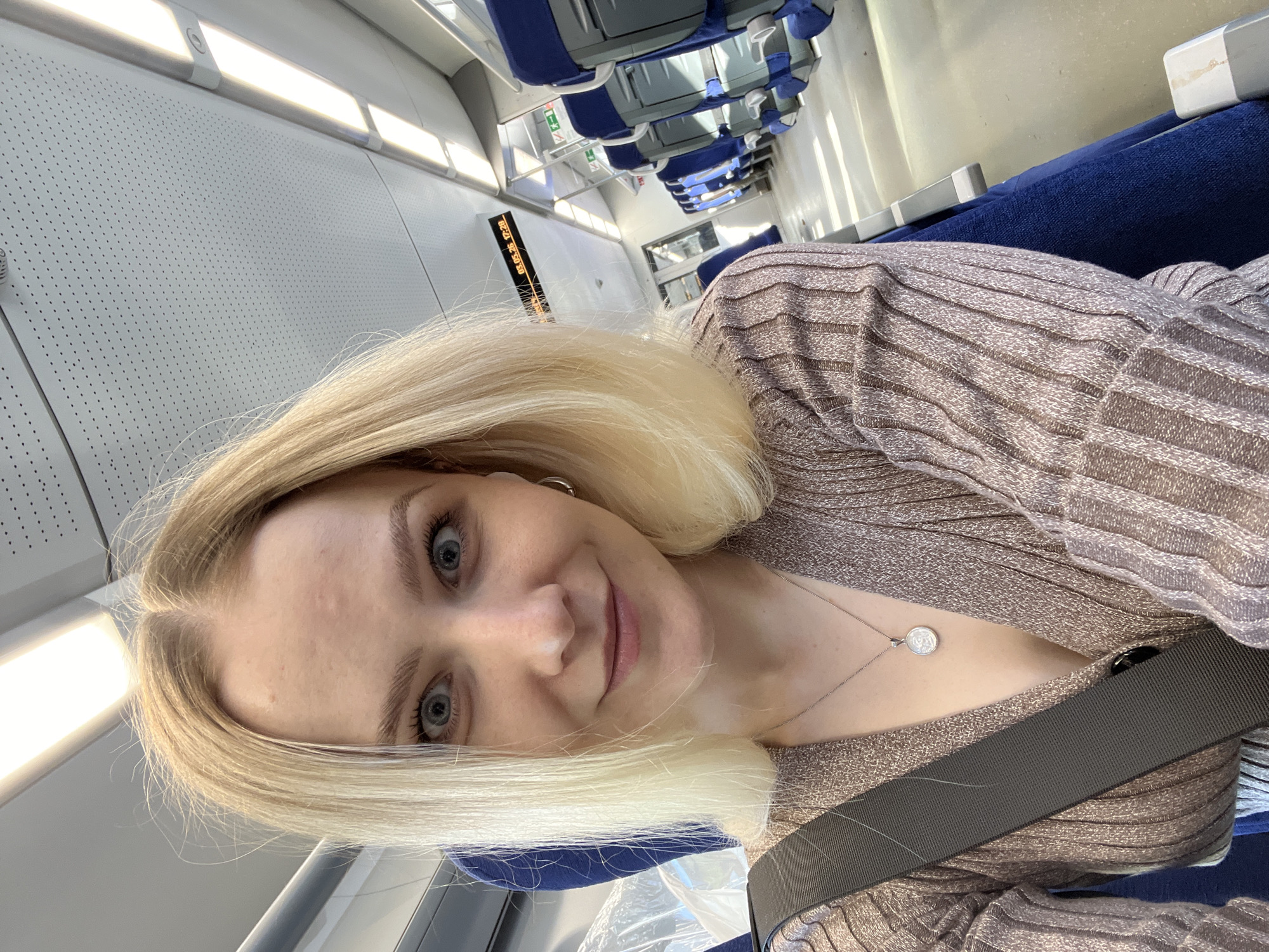 A blonde woman is taking a selfie on a train. She is wearing a silver necklace with a round pendant and a bronze colored, striped sweater. She is wearing a seatbelt. Behind her are rows of blue train seats. The train's interior is gray with small, repeating holes. The train appears to be moving. There is a digital sign showing a schedule on the train wall. There is no indication in the image of the city of Khimki, Russia.