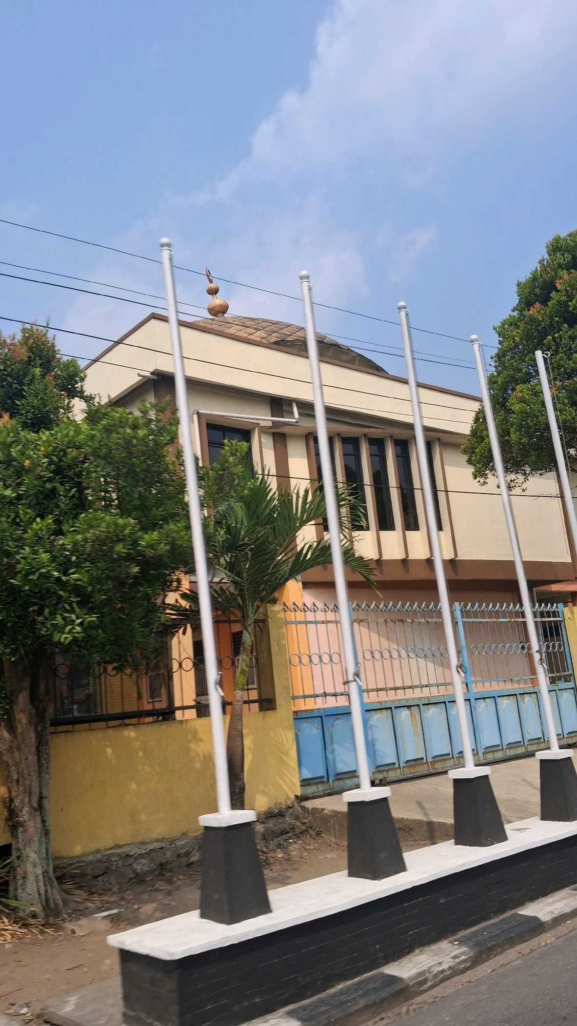 A low-angle outdoor shot captures a building with a tiled roof and a small dome structure, possibly indicating a mosque, in Ambartawang, Indonesia. The building has a cream-colored facade with brown accents and dark-framed windows. In front of the building, a row of five flagpoles stands on concrete bases, with two flagpoles cut off by the frame. A fence, partly painted blue and orange with an intricate metal design, separates the building from the foreground. Lush green trees are visible to the left and right of the building, and a palm tree stands in front of the fence. The sky above is clear blue with scattered white clouds, and electrical wires are strung across the upper portion of the frame. The foreground shows a paved surface, possibly a road or sidewalk, with a black and white striped edge.