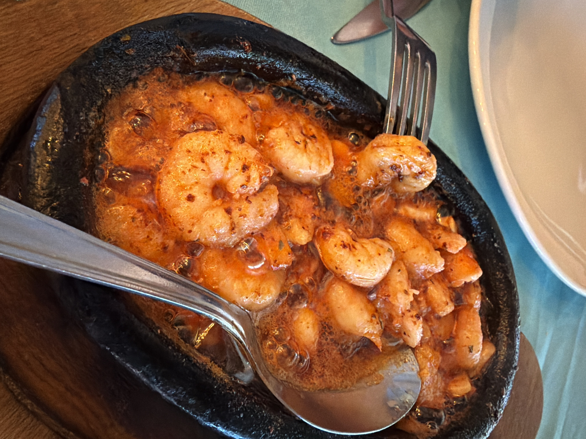 This is a close-up, overhead shot of a sizzling hot pan of shrimp served in a dark, oval-shaped dish. The shrimp are coated in a vibrant orange, oily sauce with visible spices. A silver spoon is nestled in the dish, scooping up some of the shrimp and sauce. A fork is visible to the left of the dish, with one tine touching a shrimp. The dish is presented on a wooden board. In the background, a white plate and a turquoise tablecloth are partially visible, suggesting a restaurant setting. The image captures a moment of a meal being served or enjoyed. There are no people visible in the image.