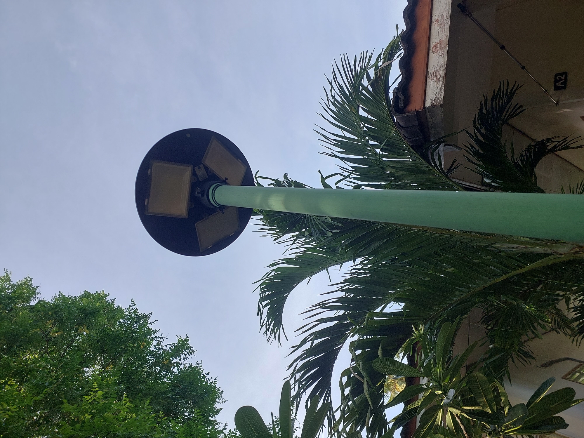 An outdoor, low-angle shot captures a tall, green lamppost in Baan Mae Nam, Thailand. The lamppost is topped with a large, circular fixture containing four square light panels. Palm fronds and other tropical foliage surround the base of the lamppost, with the roof of a building visible behind them. The sky above is a pale blue with faint clouds.