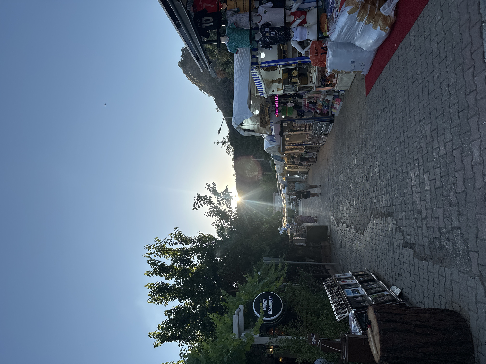 The image captures a sunlit street scene in Fethiye, Turkey. The perspective is along a pedestrian walkway paved with gray stones, extending towards a bright setting sun obscured slightly by trees and a hillside. On the right, vendor stalls display a variety of merchandise including clothing (sports jerseys), souvenirs, and bags. Several people are walking in the distance. At the lower right, a display of leather art sits alongside a tree stump. A round sign for "Mimoza Restaurant" is visible on the left side. The sky is a clear blue, with a small bird visible.