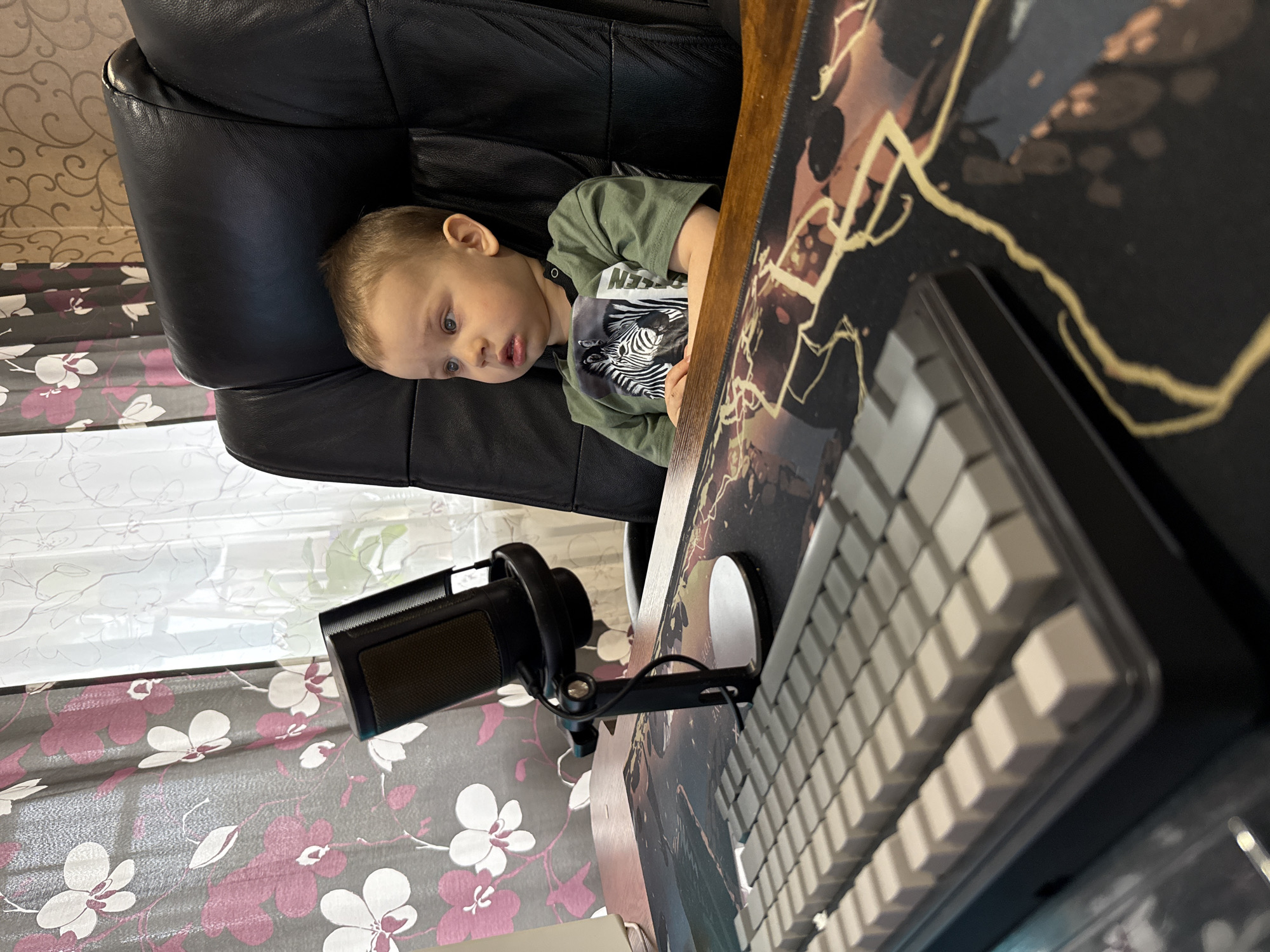 A young child, likely a toddler or preschooler, lies on a black leather armchair, looking intently towards a desk in an indoor setting in Заволжье, Russia. The child has light brown hair and blue-grey eyes, with a neutral to slightly curious expression. They are wearing a green short-sleeved t-shirt featuring a graphic of a zebra head and the partial word "EEN" printed in white text.
To the right, a wooden desk is partially visible, covered by a large dark mousepad with an abstract design of light gold lines and brown/orange shapes. A mechanical keyboard with numerous light-colored (off-white/grey) keycaps is placed on the mousepad. A black studio microphone, equipped with a pop filter and mounted on an articulating arm, is clamped to the desk's edge.
In the background, behind the child and chair, are windows adorned with two layers of curtains. The inner layer is sheer white with a subtle floral pattern, while the outer layer is grey with distinct purple and white floral motifs. A light-colored wall with a faint patterned wallpaper is also visible behind the armchair. The natural, bright lighting suggests it is daytime.