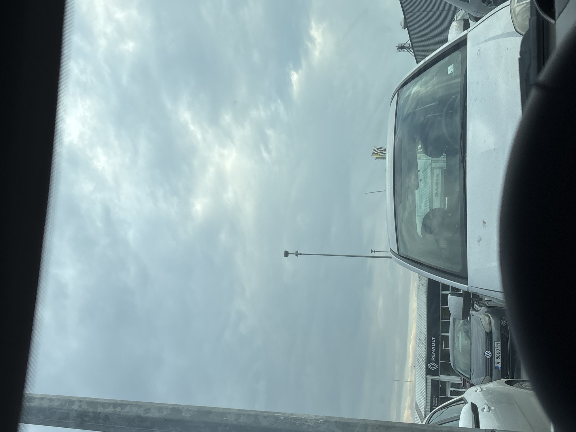 The image, taken from inside a vehicle looking out, shows a cloudy sky above a car dealership or parking lot. The dealership appears to have Renault and Dacia vehicles. A gray Volkswagen Up! is visible, bearing a Bulgarian license plate "A 8660 HH." The location is plausibly Burgas, Bulgaria, given the brands of cars and license plate format.