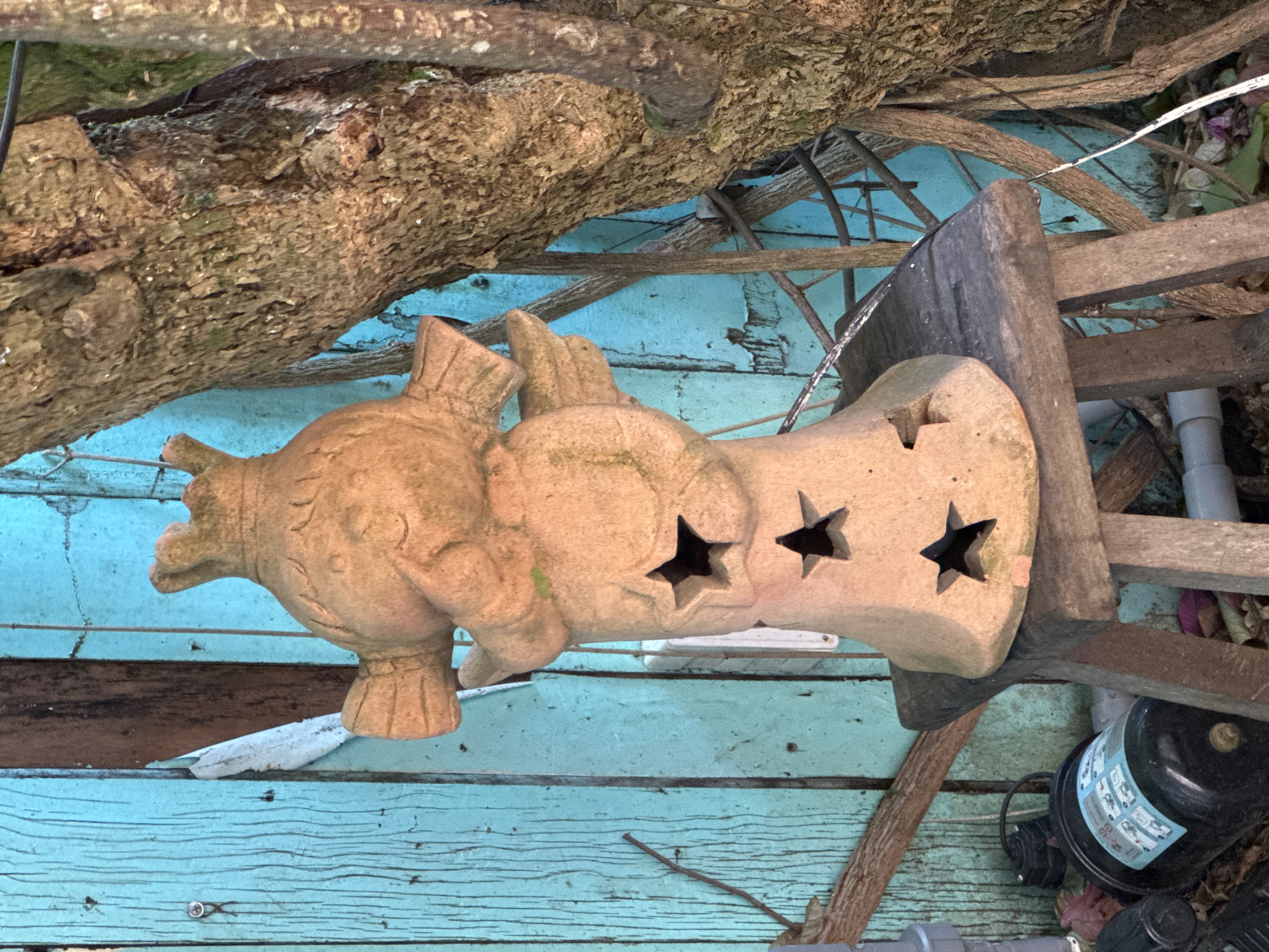 A terracotta statue of a character with a crown and wings stands on a wooden stool in front of a weathered, turquoise wooden fence in Da Lat, Vietnam. The statue has cutouts in the shape of stars on its lower body. To the right of the statue, a tree trunk with rough bark and tangled branches is visible. In the foreground, to the left of the stool, a black cylindrical object with a blue label and some plumbing pipes are on the ground. The ground is covered with fallen leaves and debris. The scene appears to be in an outdoor, possibly garden, setting.