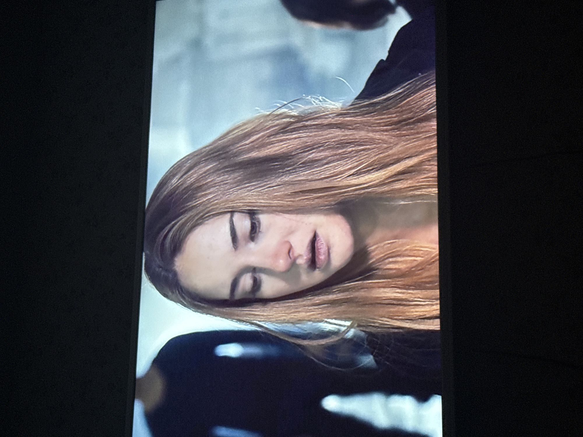 A close-up shot depicts a woman with light brown hair, looking downward with her eyes partially closed and mouth slightly open. The background is blurry and suggests an indoor setting with a muted color palette. Fragments of other figures appear blurred in the periphery. The photograph was taken at an angle, likely capturing a screen display. I have no information about the city of Mytishchi, Russia, or its connection to this photo.
