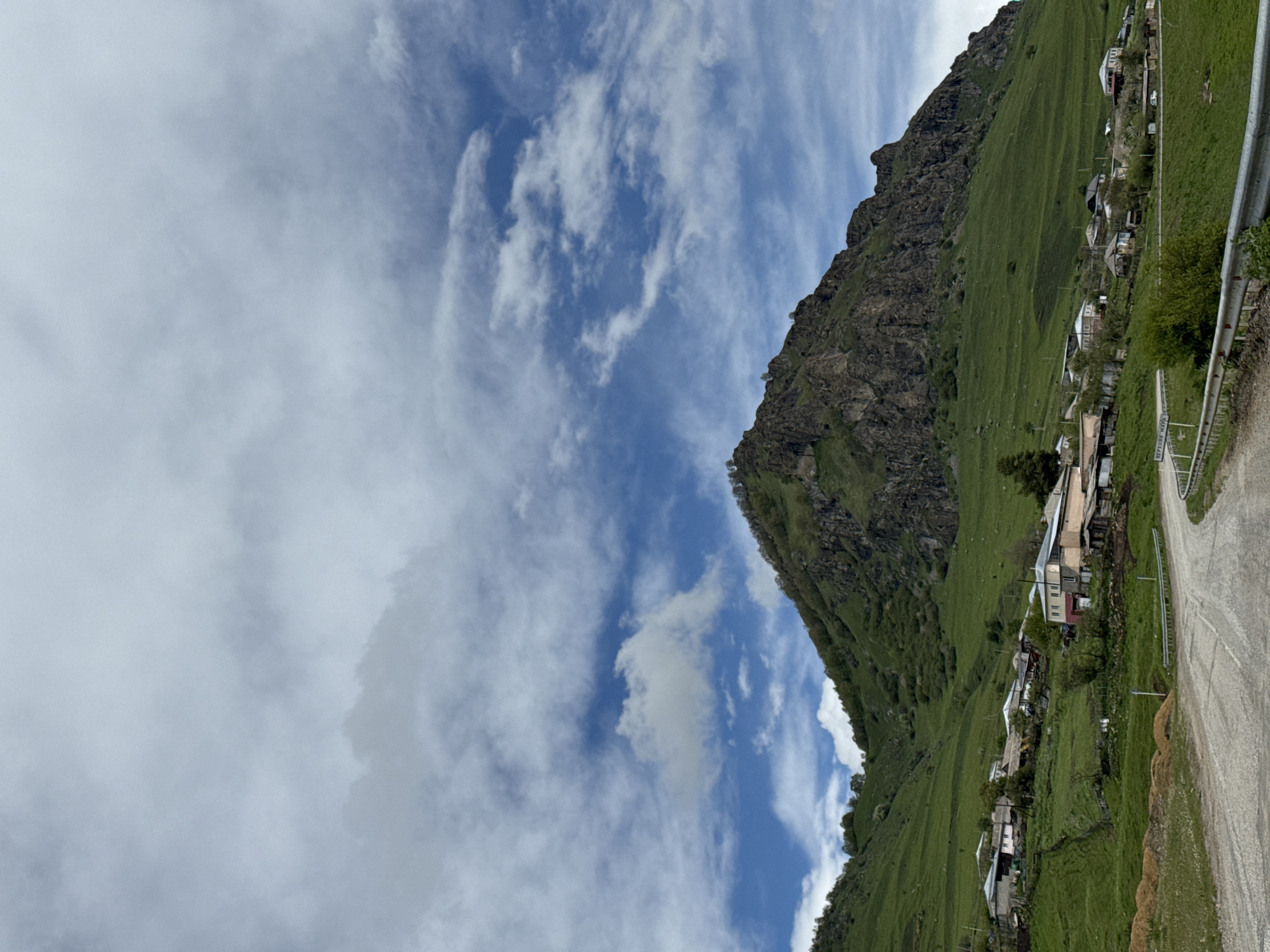 The image shows a landscape in Нижняя Мара, Russia, featuring a mountain with a rugged, rocky top and grassy slopes. A village is situated along the slope of the mountain, consisting of houses and a road. A metal guardrail is visible along the road. The sky is blue with scattered white clouds.