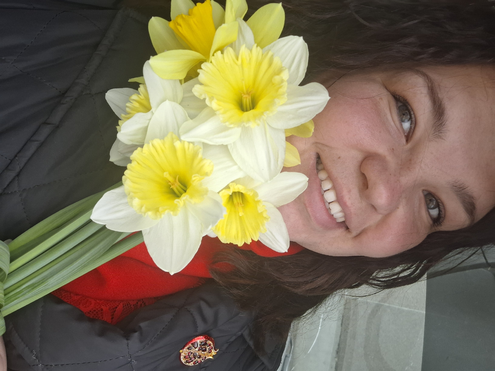 A smiling woman holds a bouquet of yellow and white daffodils. She wears a black, quilted jacket with a red scarf peeking out at the neckline. A decorative pin is visible on the jacket. The location is unclear and not definitively identifiable as Марковский, Russia.