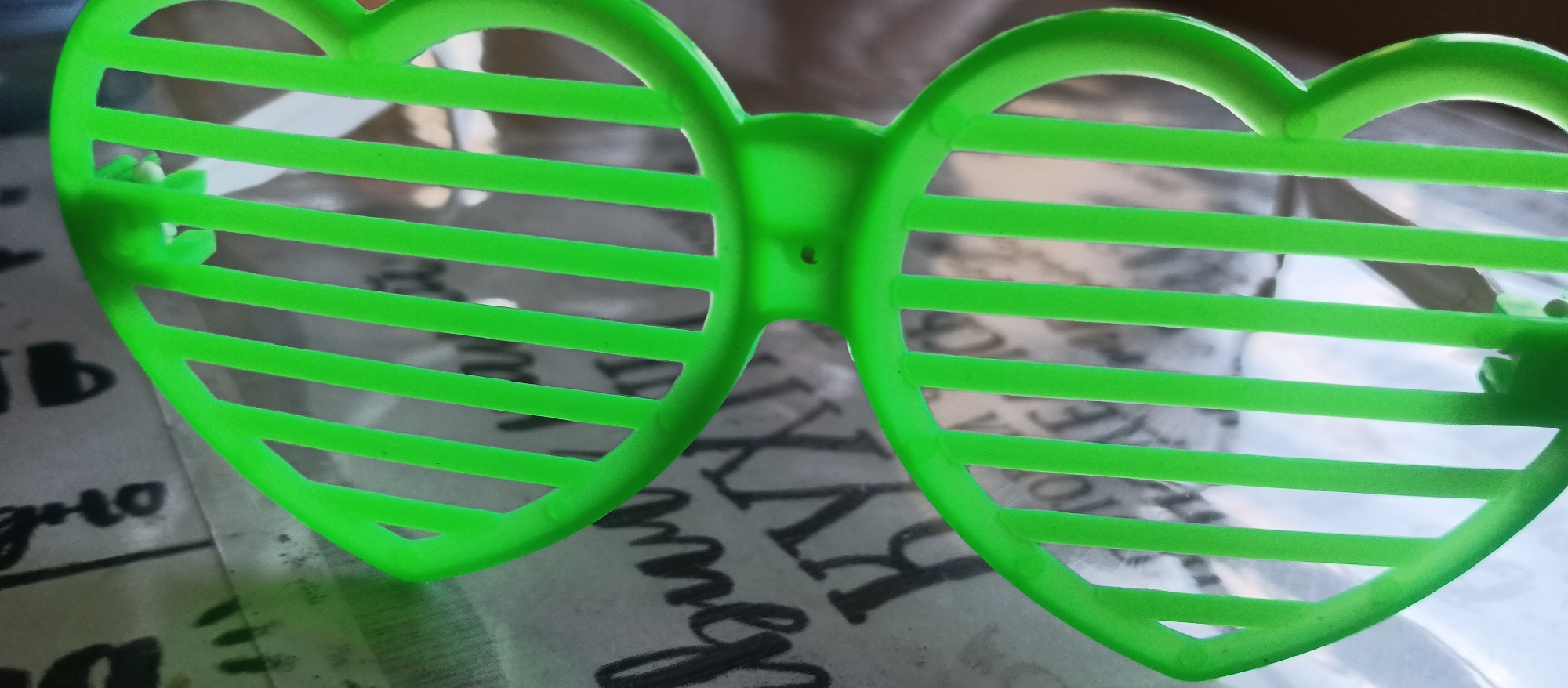 A pair of bright neon green, heart-shaped shutter shade sunglasses are prominently featured in the image. The sunglasses are made of plastic, with the frames forming two distinct heart shapes. Instead of traditional lenses, each heart-shaped opening contains several horizontal slats, typical of novelty shutter shades. A small, circular hinge connecting the two heart frames is visible at the bridge. Another hinge point for the arm of the sunglasses can be seen on the left frame.
The sunglasses are resting on a surface that forms the out-of-focus background. This background appears to be patterned with stylized Cyrillic text. Visible letters and fragments include "Ь", "ъ", "Я", "М", "Н", and partial words like "дно". The text's decorative style suggests it might be on a tablecloth, wrapping paper, or another textured surface rather than plain print.
The image captures the sunglasses in a static position, with no discernible activity taking place. The lighting suggests an indoor environment. No specific time of day or emotional context can be inferred.