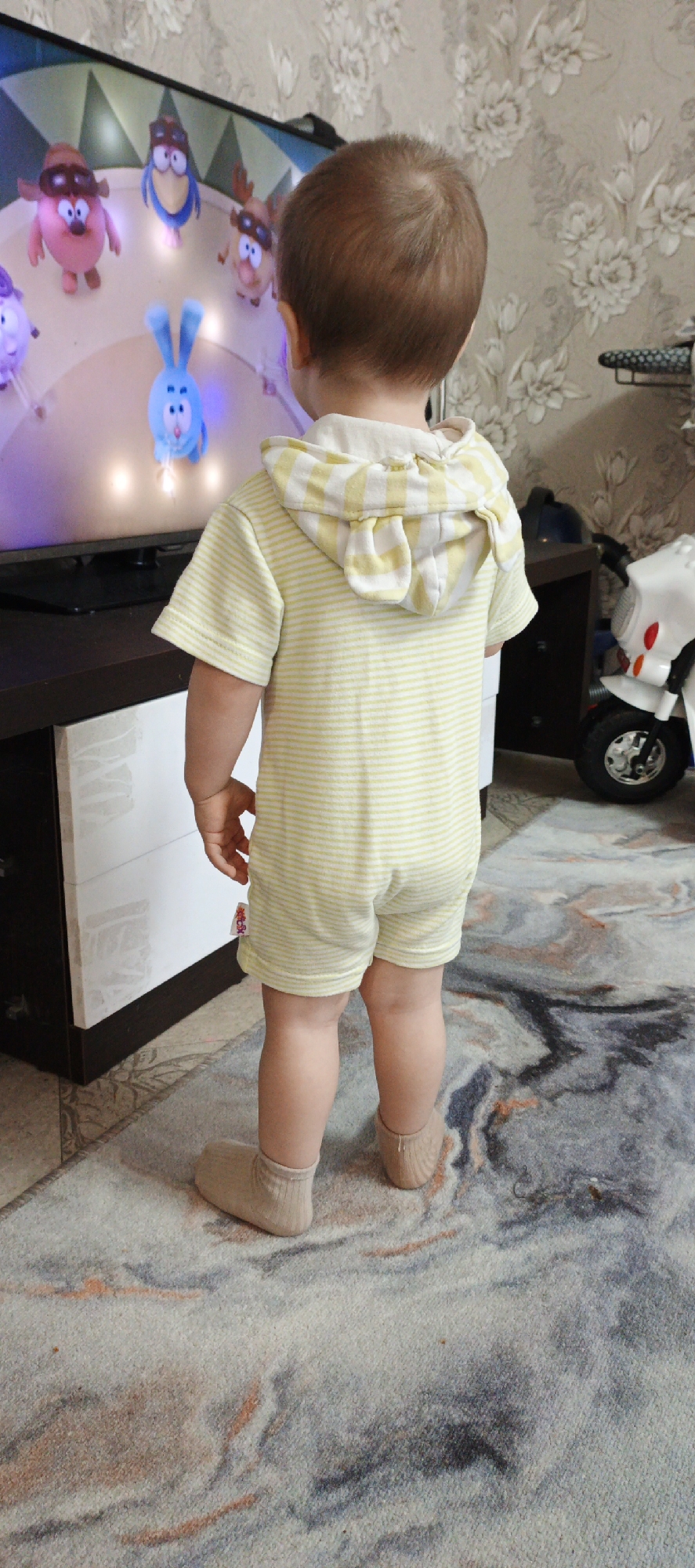 A young child, seen from behind, stands facing a television screen, engrossed in watching a cartoon. The child is wearing a light yellow and white striped, short-sleeved hooded romper that has small ear-like features on the hood, and light-colored socks. Their attention is solely directed towards the TV, which displays several colorful animated characters from the Russian series "Kikoriki" (Smeshariki). Visible characters include a blue rabbit (Krosh), a pink pig (Nyusha), a blue crow (Kar-Karych), and a brown moose (Losyash), arranged in a circular pattern on a bright background.
The television rests on a dark brown entertainment unit with white cabinet doors. To the right of the child, a white and red ride-on toy, resembling a small motorcycle, is partially visible. The floor is covered by a patterned rug featuring abstract designs in shades of grey, blue, and hints of orange/brown. The wall behind the TV and child is adorned with wallpaper displaying a subtle floral pattern in light tones. The scene takes place indoors; the time of day and external weather are not discernible. A small red tag is visible on the child's romper, but any text on it is illegible.