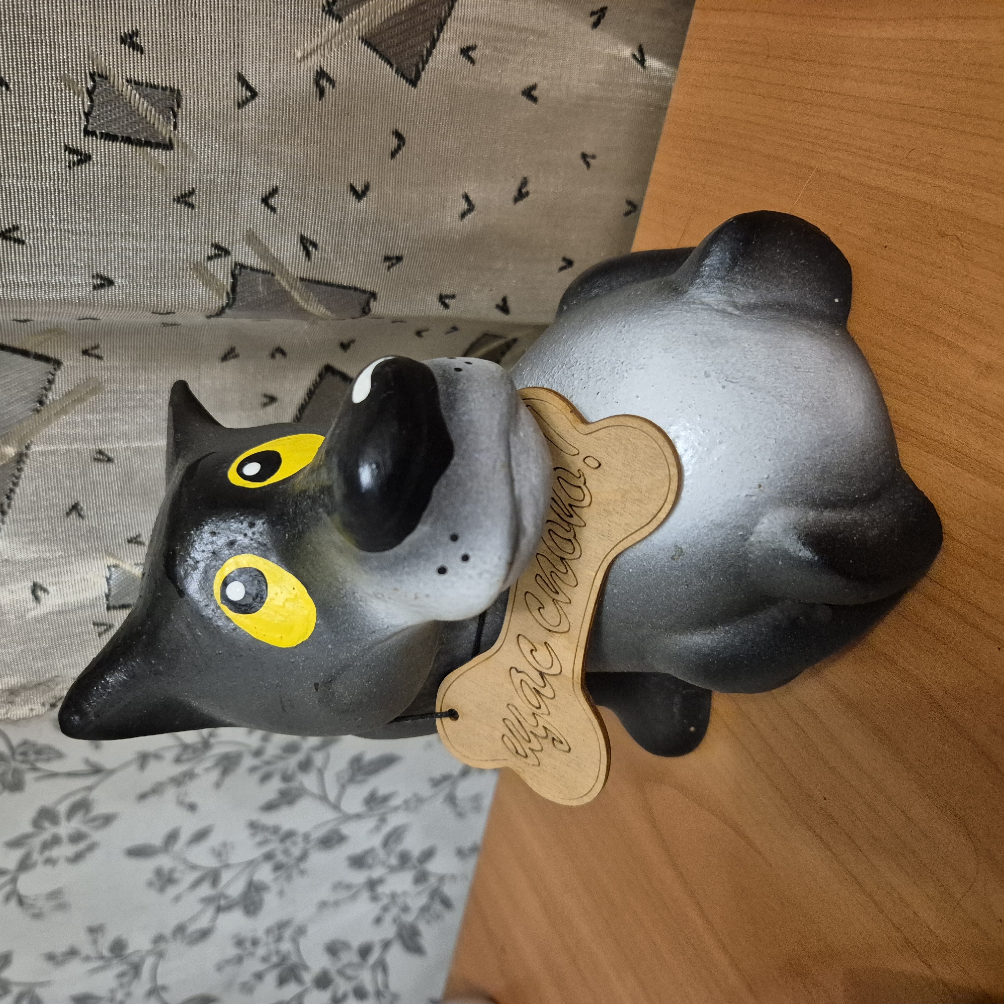 A ceramic dog figurine, painted black and gray with bright yellow eyes and a white tip on its snout, sits on a wooden surface. A bone-shaped wooden tag is suspended around its neck by a black string, with cursive text engraved on it. The background features a floral patterned curtain and another curtain with a geometric design. The scene is indoors, likely a room within a residential building in the city of Liski, Russia. There are no people present in the image.