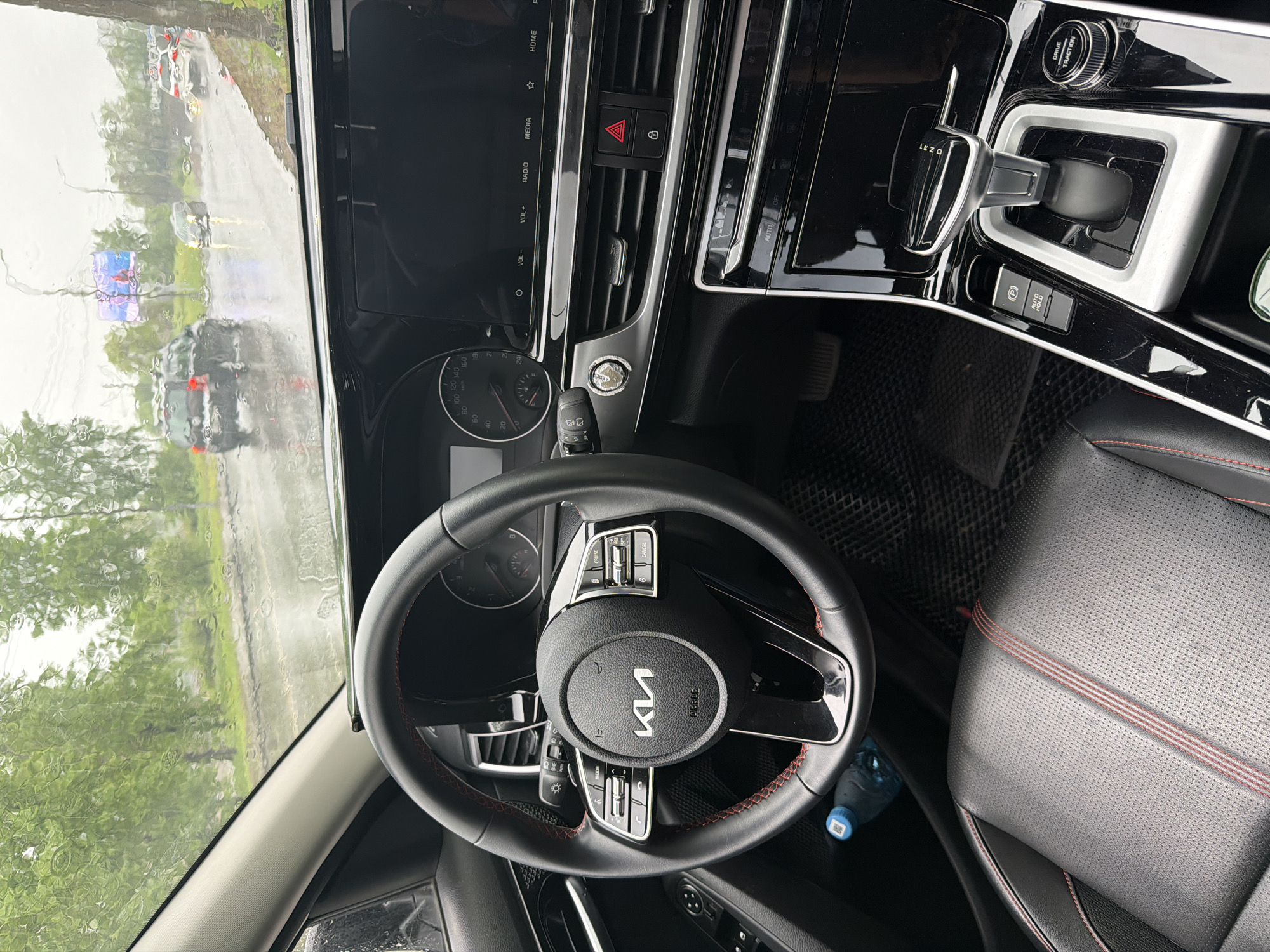The image depicts the interior of a Kia vehicle, specifically the steering wheel and dashboard area. The Kia logo is prominently displayed in the center of the steering wheel. The dashboard includes a digital display screen, air vents, hazard light button, and a push-button start. The gear shift is visible, and a "Drive/Traction" knob is located nearby. The seat is black leather with red stitching. A water bottle is visible on the floor.
Outside the car window, it appears to be raining, with blurred images of trees, roads, and other vehicles visible. Based on the Russian script on the steering wheel (Ки), it is likely the car is in Russia, and the additional OCR text (DRIVE/TRACTION) is written in English. There is insufficient information to confirm or deny that this image was taken in Krasnoyarsk, Russia.