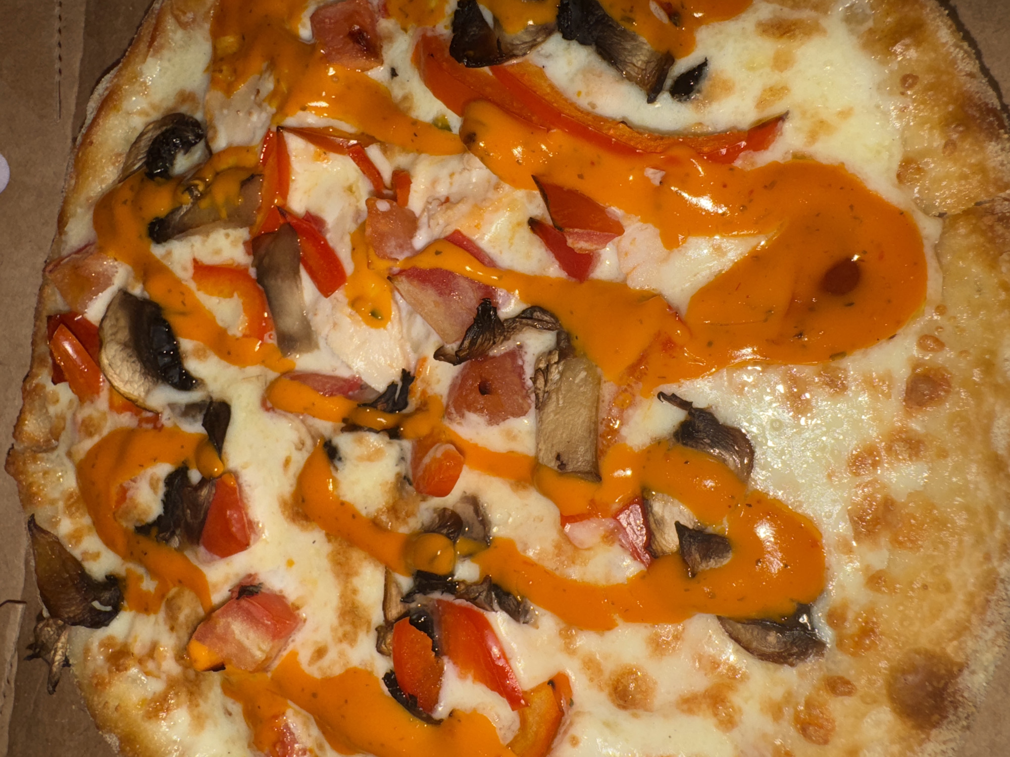 This image is a close-up, overhead shot of a pizza in a cardboard box. The pizza has a golden-brown crust and is topped with melted white cheese, sliced mushrooms, and diced red bell peppers. A vibrant orange sauce, possibly a spicy aioli or sriracha-based sauce, has been drizzled generously over the toppings in a decorative pattern. The pizza appears to be freshly made and is the sole subject of the image. The background is a blurry, brown cardboard box, suggesting the pizza is either being served or is in transit. There is no information in the image to suggest it is located in Chaikovsky, Russia, or any specific city.