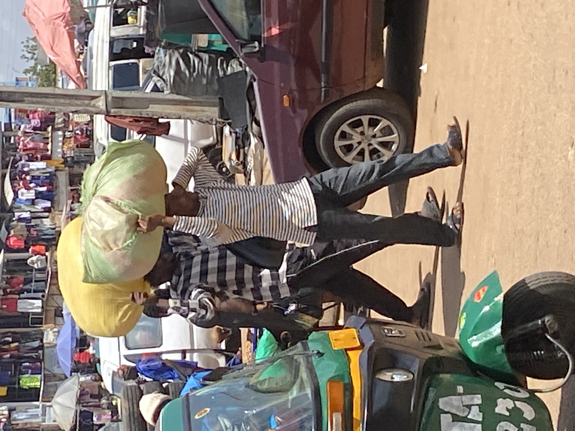 This image depicts a bustling street or market scene in Bukuru, Nigeria, captured during bright daylight. The unpaved ground suggests a dirt or gravel pathway.
In the foreground, two men are prominently featured, both engaged in the common practice of head-carrying large sacks. The man in front wears a light-colored shirt with dark vertical stripes and dark trousers, along with sandals. He balances a large, light-green sack on his head. Behind him, another man, clad in a checkered long-sleeved shirt and dark trousers, carries a substantial yellow sack on his head, possibly supporting the green one above.
The left side of the frame reveals a vibrant market area with numerous stalls displaying various goods. Several open umbrellas, one light blue and another white, offer shade over these market stands. On the right side, vehicles are visible. A dark red or maroon car with alloy wheels is parked or moving. In the immediate foreground on the right, the front portion of a green and black auto-rickshaw is seen, showing its windshield and a headlight. Partially obscured white text, including fragmented letters such as 'E', 'G', and a number like '4' or '74', is visible on its side. Indistinct red text can also be faintly observed on a background sign. The overall scene portrays lively commercial activity and daily life in an active, urban or semi-urban environment.