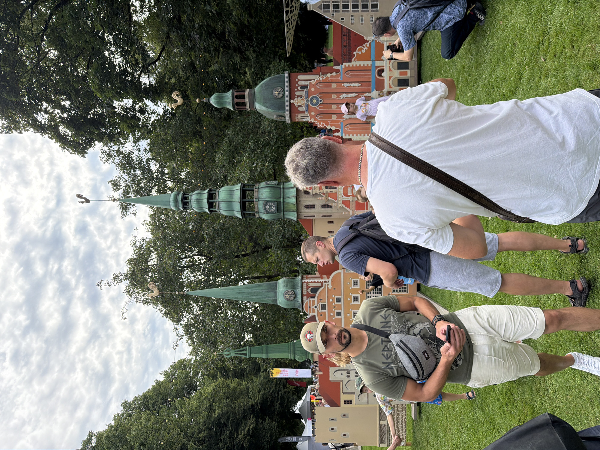 The image depicts an outdoor scene in a park-like setting, likely in Riga, Latvia, given the presence of a miniature replica of the Riga skyline, featuring iconic spires and buildings. The sky is partly cloudy. In the foreground, a man wearing a green t-shirt, light-colored shorts, a baseball cap, and a crossbody bag is looking towards the camera. To his right, another man in a dark t-shirt and grey shorts is visible from the side. In the background, the miniature city model is prominent, with several people interacting with it. One person is taking a photo of the model. The ground is covered in green grass, and trees form a backdrop to the scene.