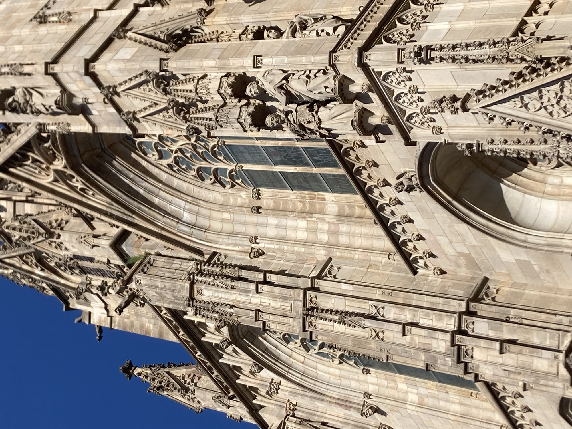 The image shows a close-up, low-angle view of the exterior of the Barcelona Cathedral in Barcelona, Spain. The gothic architecture is characterized by intricate stonework, pointed arches, and decorative carvings. Several gargoyles are visible on the upper portions of the building, and sculptures of figures in robes are mounted on ledges. The building is constructed of light-colored stone, and the sky above is a clear, deep blue. The image captures the detail and grandeur of the cathedral's facade, with shadows playing across the stone due to the sunlight.