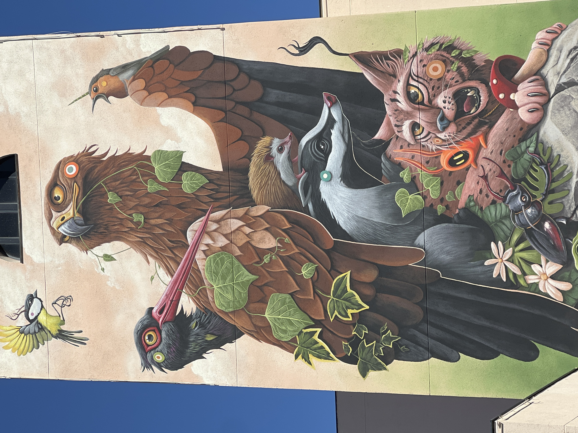 This is a photograph of a mural on a building in Madrid, Spain. The mural depicts a collage of animals. From left to right are: A yellow and black bird, 3 birds of prey covered in green leafy vines, a hedgehog, a badger, a wildcat, a mushroom, a pair of hands, and a large beetle with white flowers.