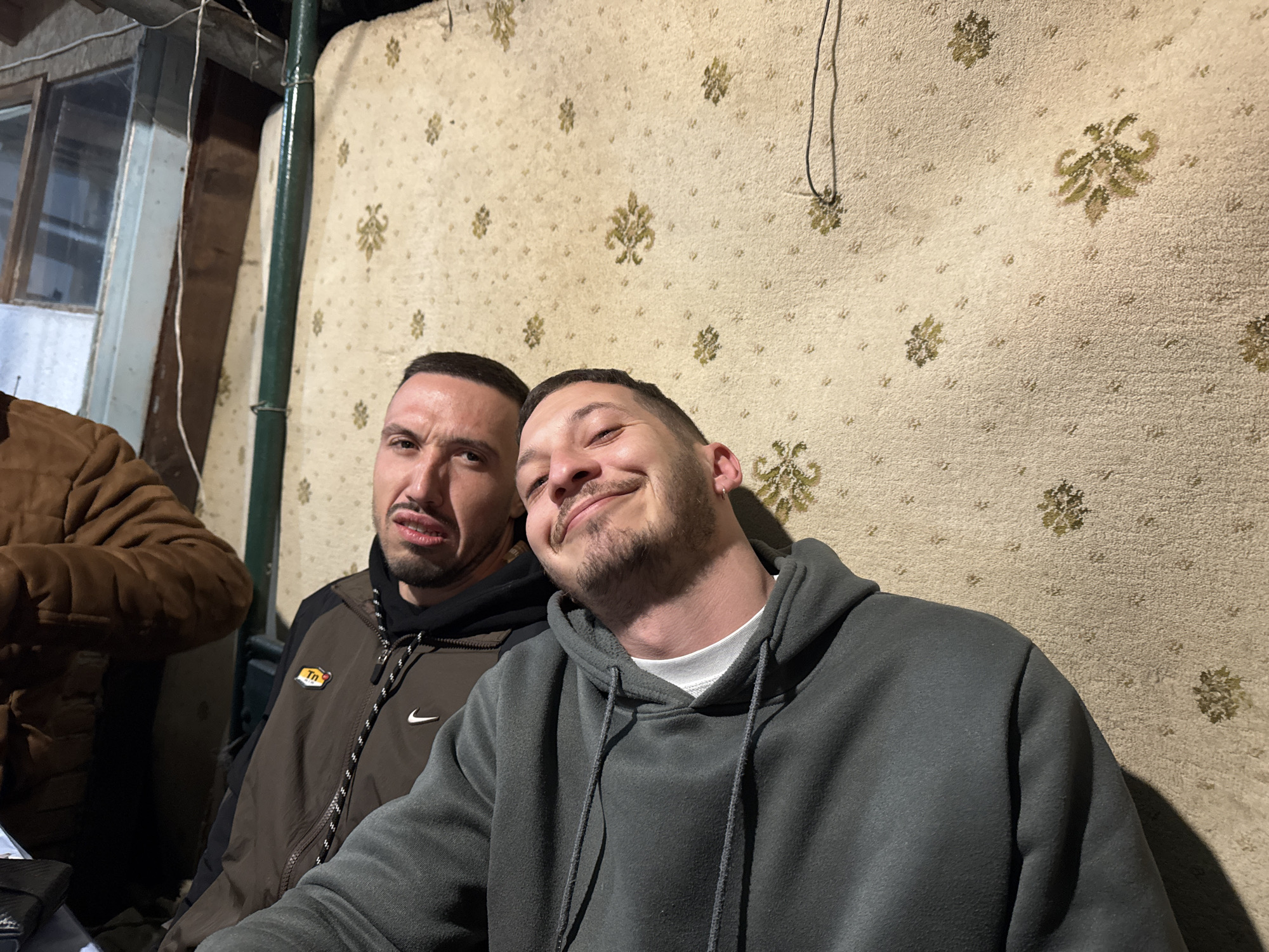 Two men are sitting close together indoors, with one leaning his head towards the other. The man on the right wears a gray hoodie with the hood down, a white t-shirt is seen beneath. The man on the left is wearing a brown jacket with black details and a white Nike logo. Behind them is a wall covered with wallpaper featuring a pattern of repeating floral or leaf-like motifs in a light color. In the background, there's a structure that appears to be a window or a small room with glass panes. A partial view of a person wearing a brown jacket is visible on the left. The location is Kondolovo, Bulgaria.