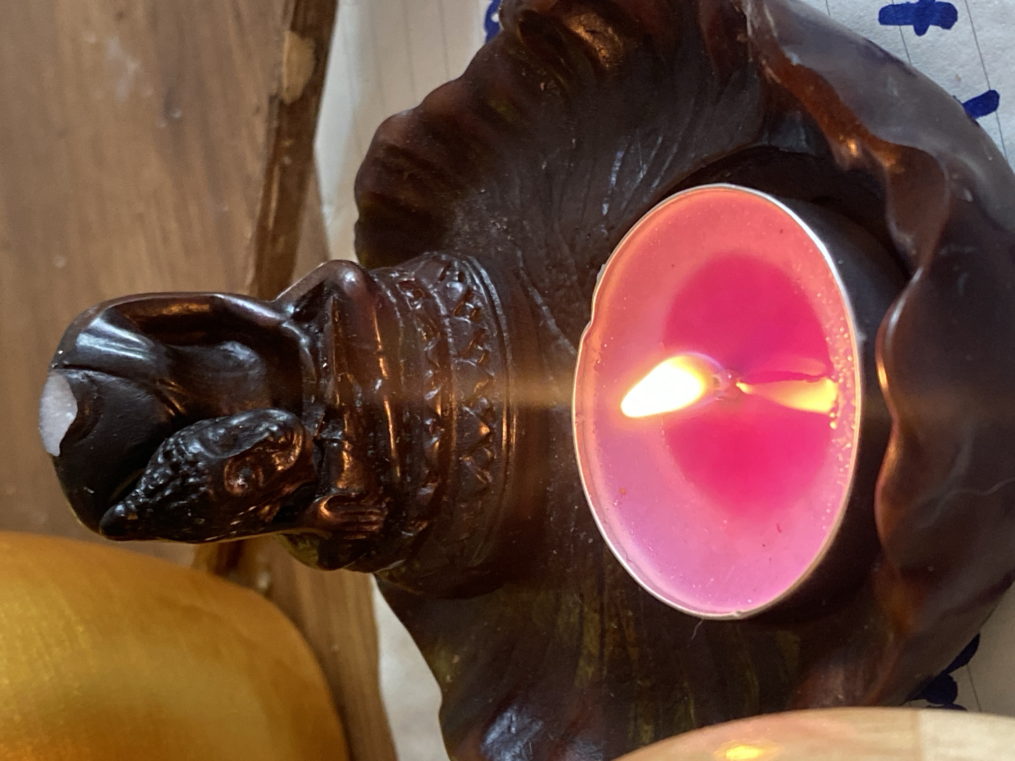 A lit pink votive candle sits in a dark, possibly wooden, carved holder. The holder appears to depict a figure, possibly a Buddha, with intricate carvings on its neck and head. The location, Baan Kai, Thailand, is provided context. The setting appears to be indoors on a wooden surface with other round objects. A blue and white patterned surface is also visible in the background.