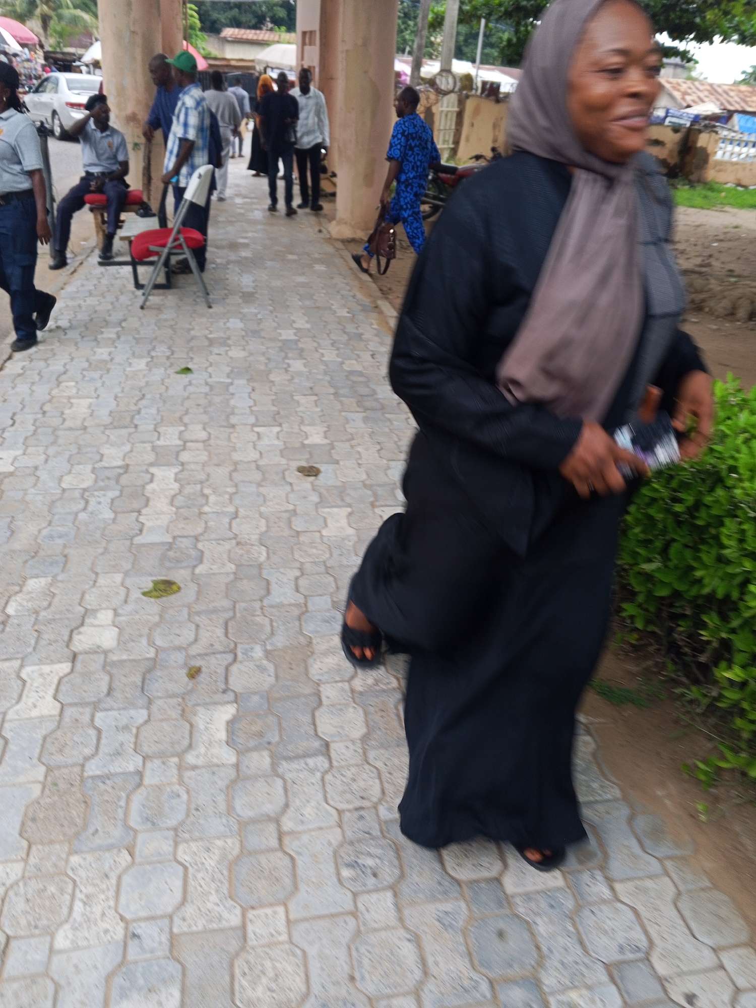 The image depicts a street scene in Lokoja, Nigeria, featuring several individuals and a paved walkway. In the foreground, a Black woman wearing a black abaya and a light brown hijab is walking towards the right of the frame, holding a mobile phone. She is wearing sandals. To her left, a man in a blue and white checkered shirt and blue trousers is standing. In the background, other people are visible, some walking, and a few are seated on red chairs. A white car is parked on the left side of the street. The environment suggests a public area with shops or buildings lining the street, indicated by structures in the background. The ground is covered with interlocking pavers.