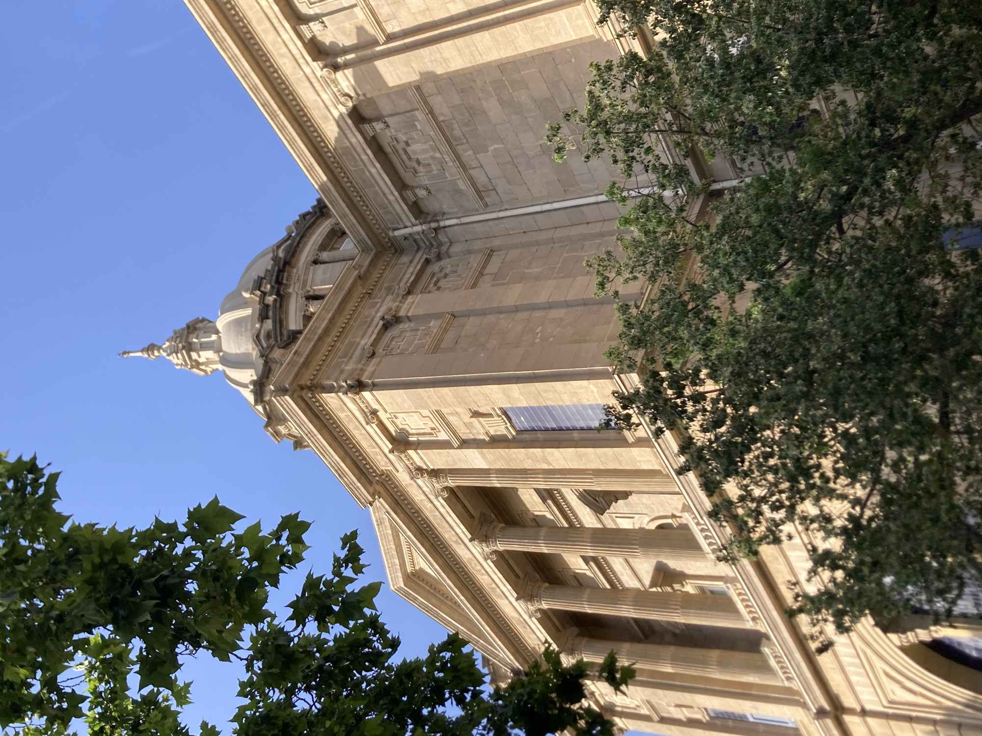 The image shows the Palau Nacional, located on Montjuïc hill in Barcelona, Spain. The building is constructed of stone and features a large dome and classical columns. Trees with green foliage are in the foreground, partially obscuring the building. The sky is clear and blue. The lighting suggests it is daytime, with sunlight illuminating the facade of the building. There are no people visible in the image.