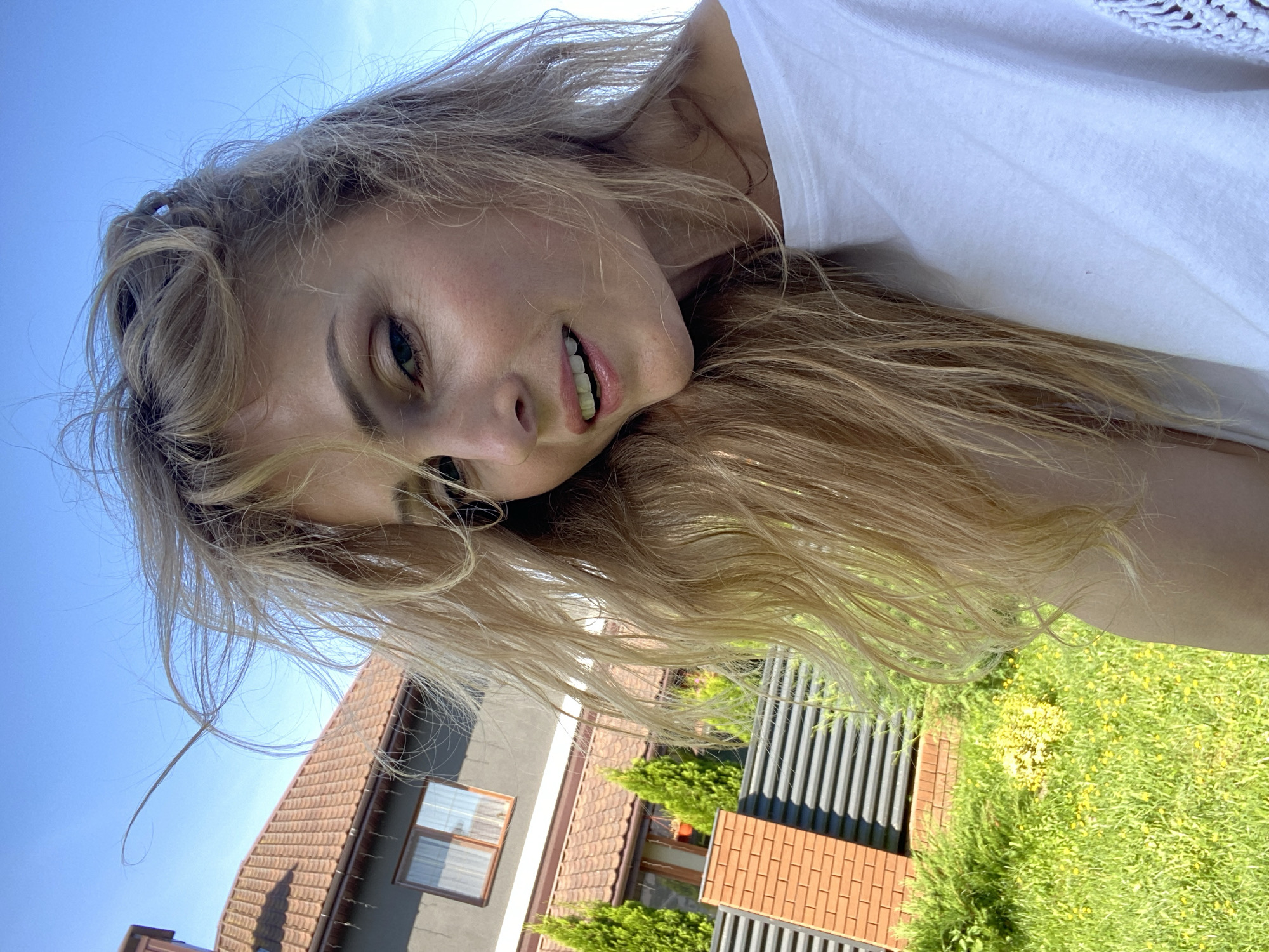 In a selfie taken outdoors, a fair-skinned woman with blonde hair styled loosely around her face smiles softly, her mouth slightly open to reveal her teeth. Her hair appears windblown, with strands falling across her face and shoulders. She is wearing a white t-shirt. In the background, a grey house with a terracotta tiled roof is visible, with windows and Christmas lights draped along the eaves. Adjacent to the house is a fence made of horizontal grey slats and vertical red brick posts. Several small, conical evergreen trees are planted behind the fence, and a patch of vibrant green grass with small yellow flowers covers the ground in the foreground. The sky above is a clear, bright blue. The scene is consistent with an outdoor setting in a residential area of Міханавіцкі сельскі Савет, Belarus, likely during daylight hours given the strong natural lighting.