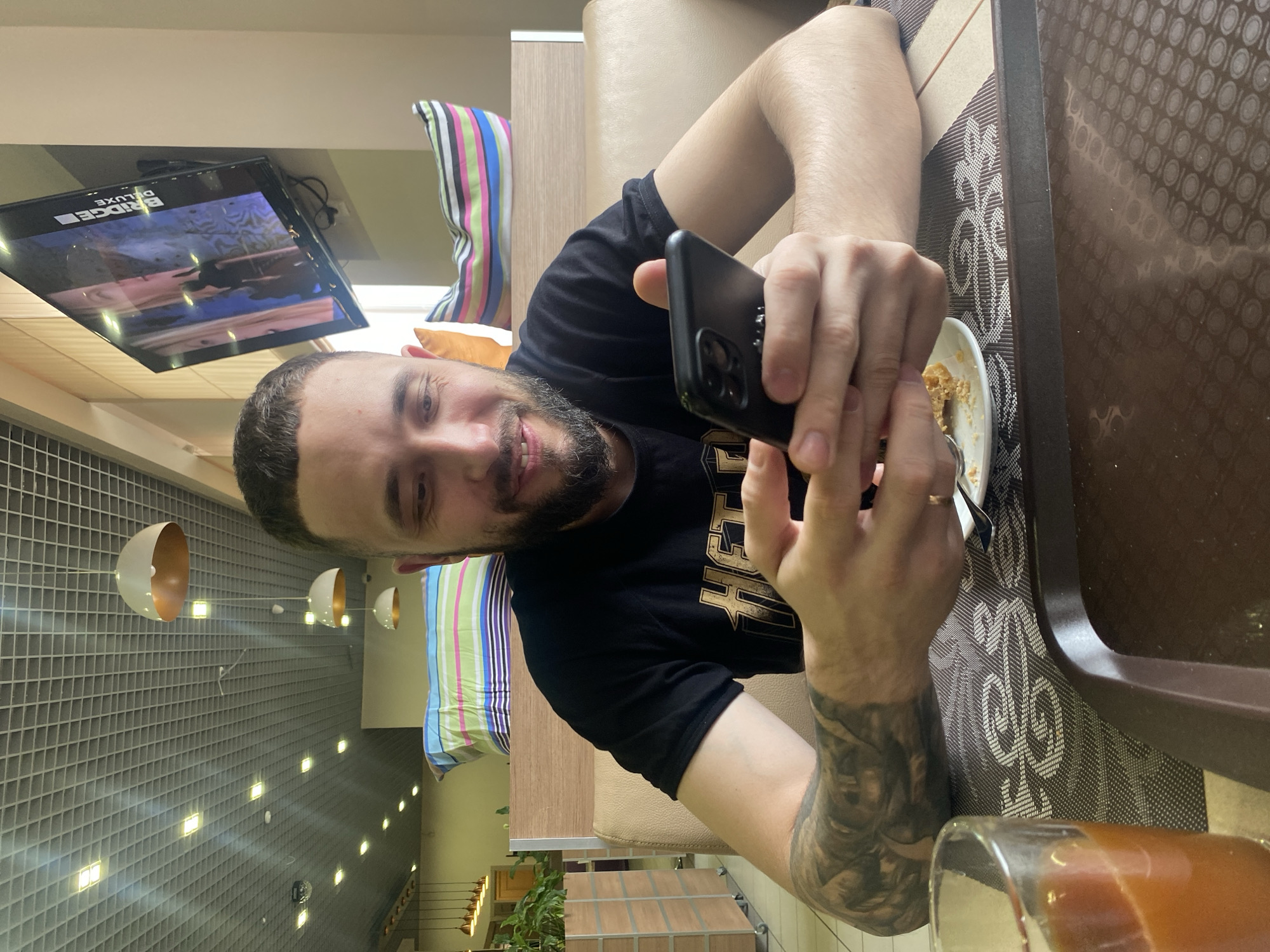 The image captures a man seated at a table in an indoor setting. He is fair-skinned, with a beard, and is wearing a black t-shirt. The man is holding a black smartphone in his hands, suggesting he might be taking a photo or video. A partially eaten dessert is visible on a plate on the table. A glass of an amber-colored liquid is in the foreground. The table has a patterned placemat. In the background, there is a television screen displaying content and a striped pillow. The ceiling has modern light fixtures, and there are plants visible in the distance. The image was taken in the city of Chaikovsky, Russia.