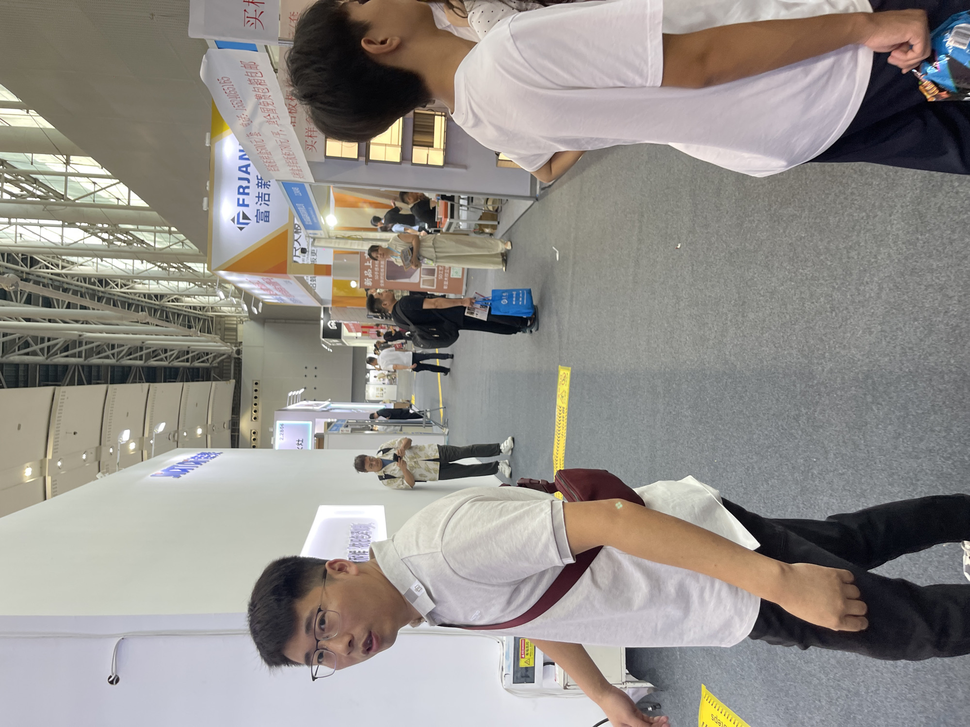 The image depicts an indoor exhibition hall in Haizhu District, China. In the foreground, a man wearing glasses, a light-colored polo shirt, and dark pants is looking to his left, with a maroon crossbody bag slung over his shoulder. His left arm is visible, showing a small, light-colored square sticker on his forearm. To his right, another person, mostly obscured from the chest down, is wearing a white t-shirt and dark pants, holding a bag of snacks in their right hand.
The background reveals a busy exhibition environment with various booths and displays. To the left, a white wall features some text and lighting. Further back, several people are walking and interacting. One man in a patterned shirt is looking at his phone. Another individual in a black t-shirt and black pants is holding a blue shopping bag. Several exhibition booths are visible, including one with a sign that reads "FRJANI" in English, accompanied by Chinese text. The ceiling of the hall shows a large industrial structure with metal beams and lighting fixtures. The floor is a grey carpeted surface.
