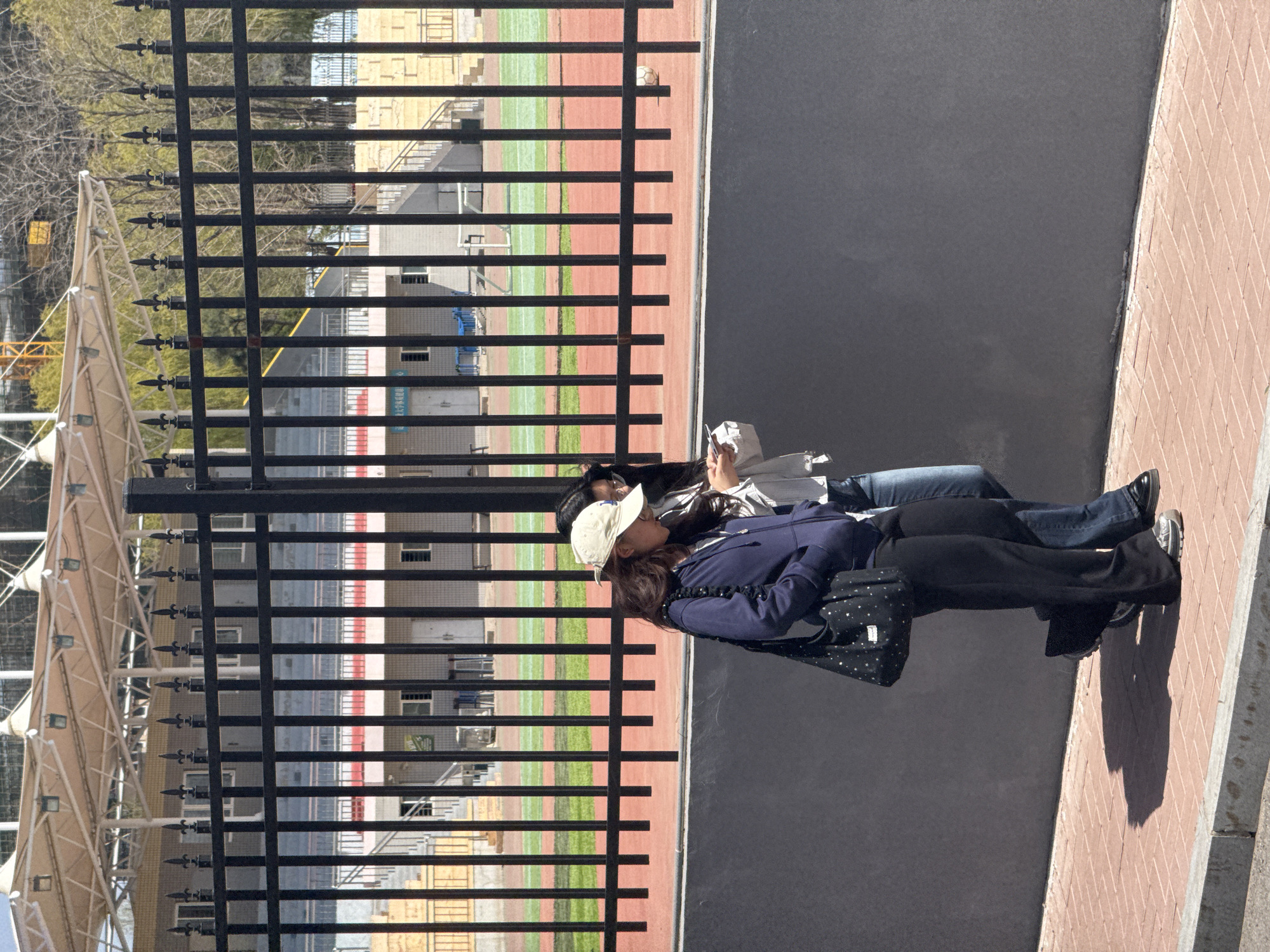 This outdoor image, taken on a bright, sunny day, depicts two individuals observed from behind a sturdy black metal fence. The fence, featuring decorative spear-top finials, runs vertically through the left half of the frame, separating the foreground from a sports field.
In the foreground, to the right of the fence, two people, likely women, are seen sitting or leaning on a dark gray paved surface next to a brick-patterned walkway. The person in front, with brown hair, wears a white baseball cap, a dark blue jacket with gathered sleeves, and black pants. She is carrying a black bag adorned with white star patterns. Partially visible behind her is another person wearing a light-colored top and blue jeans, holding what appears to be a mobile phone or small item. Their long shadows are cast on the brick walkway, indicating a high sun angle.
Beyond the fence, a vibrant green sports field with a reddish-brown running track is visible. A white soccer ball rests on the running track towards the top-right of the field. Further back, stadium bleachers and multiple buildings can be seen. Faint Chinese text is discernable on one of the buildings, possibly related to "university" (e.g., "大学") or "sports center" (e.g., "育中心"). To the far left, a large, light-colored tensile structure, possibly part of a stadium canopy or stage, stands prominently, with bare trees in the distant background. The scene suggests a relaxed atmosphere, perhaps observing an event or waiting at a sports facility in Zhongtun, China.