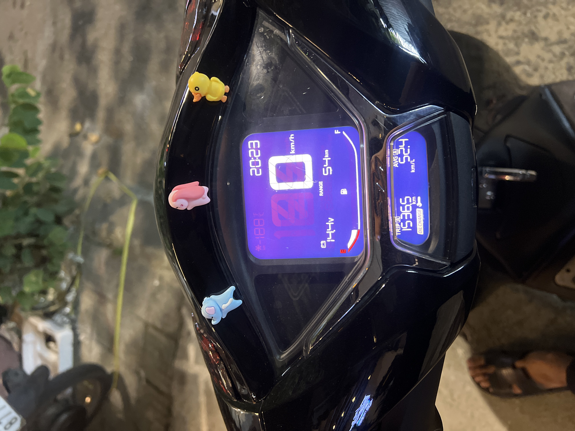 The image shows the dashboard of a motorcycle. The dashboard is black and has a digital display that shows the time as 20:23, the speedometer reading as 0 km/h, the remaining range as 54 km, the voltage as 14.4 V, and the average fuel consumption as 52.4 km/L. The odometer reads 15365 km. There are three small, decorative figures attached to the dashboard: a yellow duck, a pink pig, and a blue animal. The motorcycle appears to be parked on a street, as there is pavement visible in the background along with some greenery. A person’s feet, wearing sandals, are visible on the right side of the frame. It is not possible to confirm if the location is in Đà Nẵng, Vietnam, based on the visual information provided.