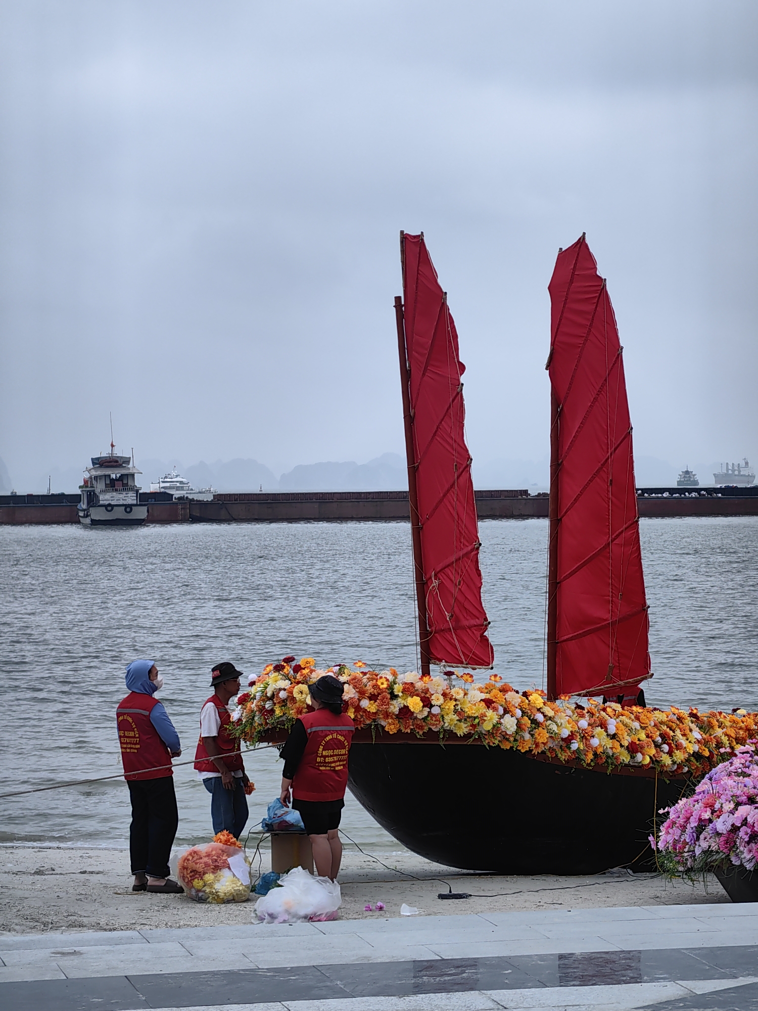 In Bai Chay Ward, Vietnam, three people are gathered near a small, black, junk-style boat decorated with orange, yellow, and white flowers along its perimeter. The boat features two red sails and is resting on a sandy shore close to the water's edge. The people are likely florists or decorators, judging by the vests they are wearing and bags of flowers on the ground. The vests have a logo and text in Vietnamese. The environment is an overcast day, and the sea has several ships and barges visible in the background. The shore is a concrete paved area which gives way to sand and water. One individual wears a blue hoodie and a face mask, while another has a black hat. The third person has a black shirt and black hat. They appear to be working on the floral arrangement on the boat.
