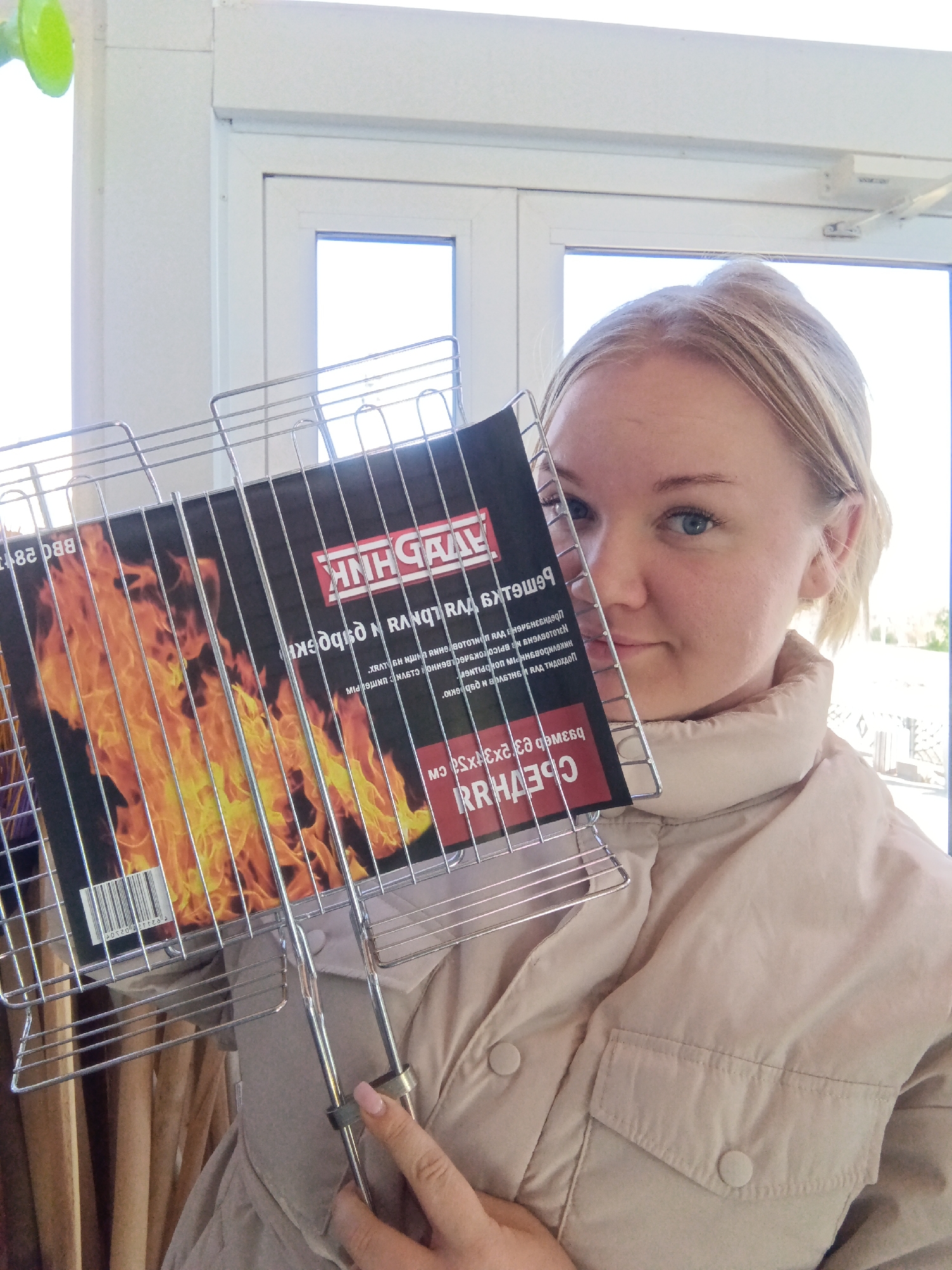 A woman holds a metal BBQ rack in front of her face indoors. The packaging on the rack depicts a fiery image. Text on the packaging includes "ANH GAAL" and upside-down Russian text. She wears a tan jacket. The background features a white-framed window and other objects in what appears to be a retail environment. There is nothing visible indicating that the photo was taken in the city of Фоки, Russia.