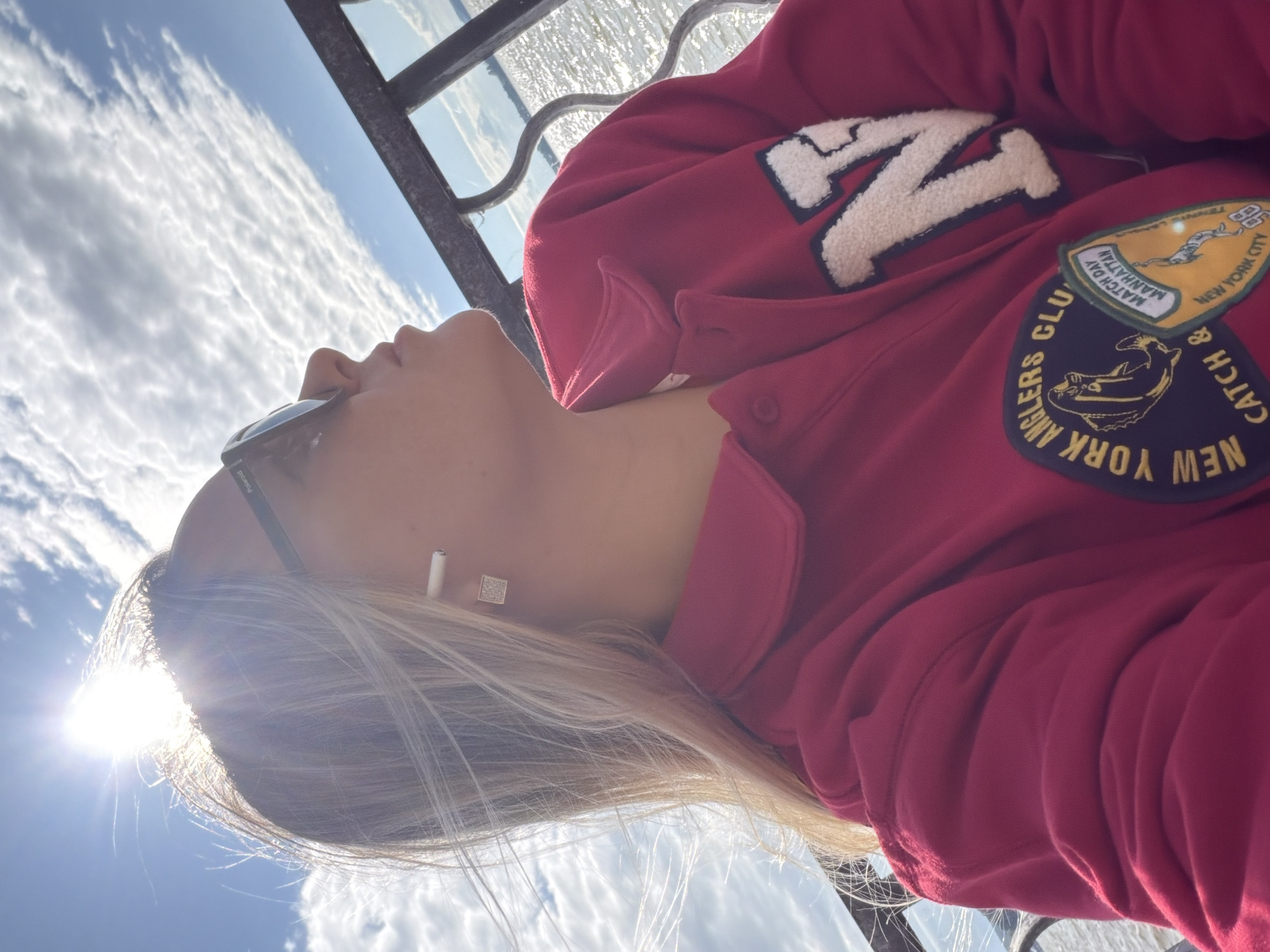 A young blonde woman with fair skin is captured in a close-up, outdoor selfie. She is wearing dark sunglasses, a bright pink jacket with an embroidered "N" and two patches, and white wireless earbuds. The sun is shining brightly, partially obscured by clouds, creating a lens flare effect. The background shows a body of water with ripples and a distant shoreline, bordered by a metal railing. The image appears to be taken on a sunny day, likely near a waterfront. There is no information to suggest the location is in Votkinsk, Russia.