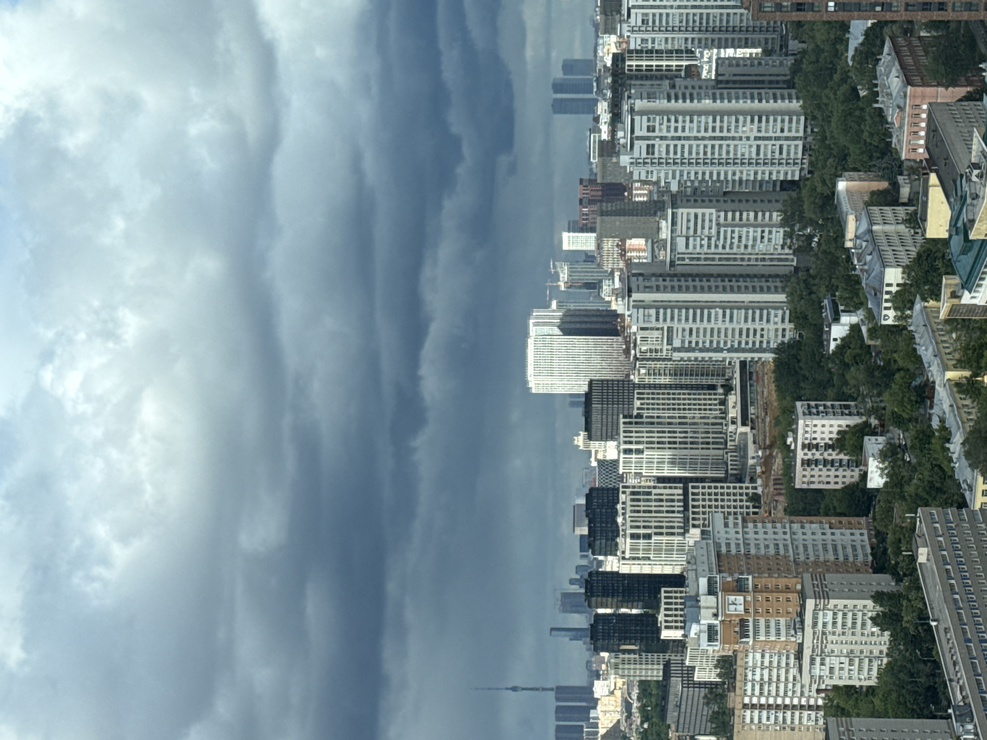 The image shows a cityscape, likely Khimki, Russia, under a sky filled with grey and white clouds. The urban landscape is characterized by high-rise buildings of varying architectural styles, interspersed with patches of green trees. A tall, thin structure, possibly a communication tower or spire, is visible in the distance. The buildings are mostly rectangular with repeating window patterns, a common design for residential or office buildings. Some structures have a lighter facade, appearing white or light grey, while others are darker, possibly due to the materials used or the effects of light and shadow.