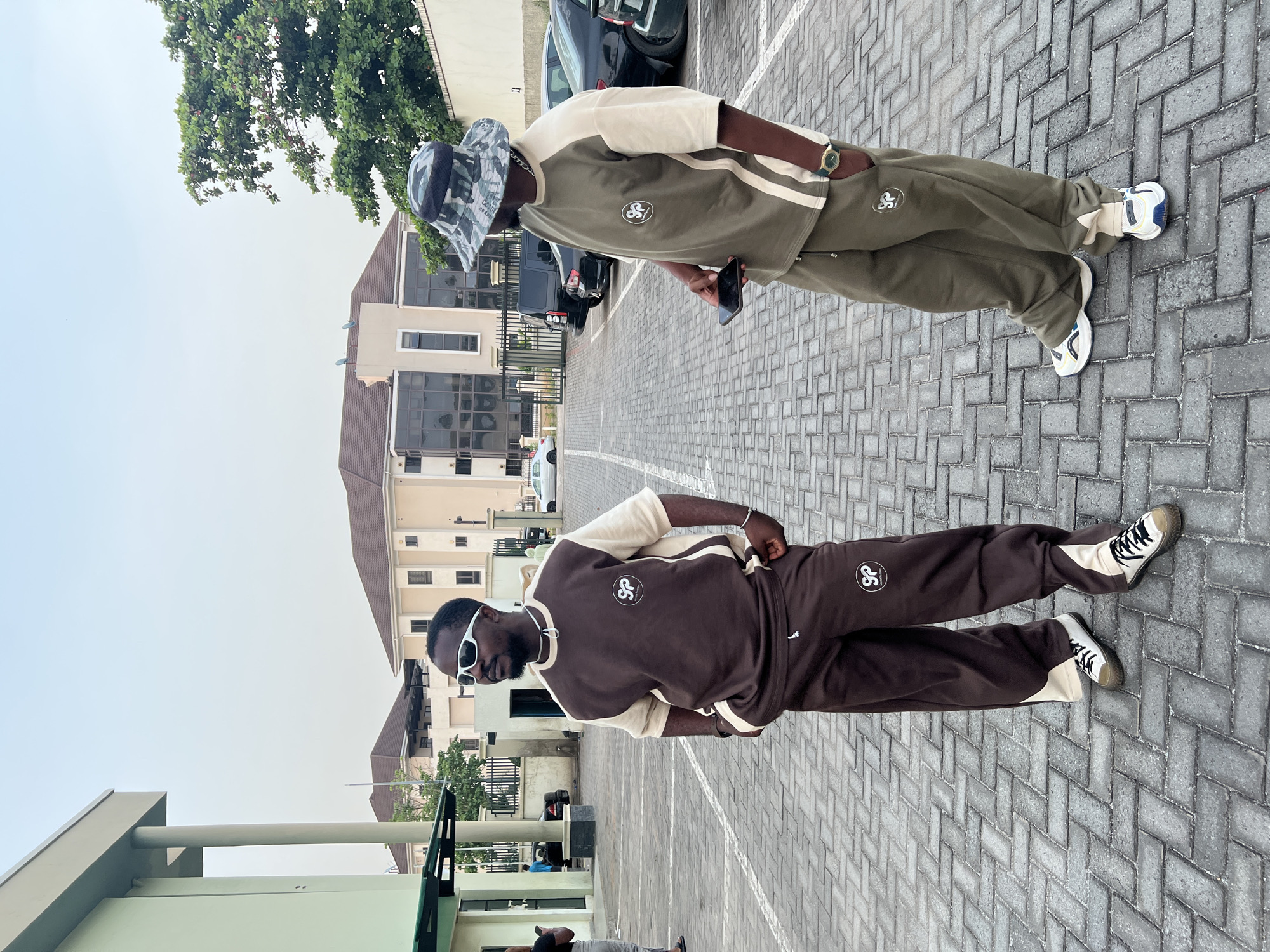 Two men are captured in an outdoor setting, likely a paved parking lot or courtyard in Abuja, Nigeria, during bright daylight hours. The ground is paved with interlocking concrete blocks. In the background, modern buildings with multiple windows and some trees are visible, along with parked cars.
The man on the left is wearing a stylish brown and beige tracksuit. Both the top and the pants feature a distinctive circular logo with the letters "SP." He accessorizes with white-framed sunglasses, a chain around his neck, and cream and black sneakers. He stands casually with one hand in his pocket.
The man on the right is dressed in a coordinated olive green and beige tracksuit, which also displays the "SP" logo on the chest and thigh. He wears a camouflage-patterned bucket hat, a gold-toned watch on his left wrist, and white and blue sneakers. He is holding a black smartphone in his right hand. Both men appear relaxed and are posing, showcasing their matching-style tracksuits.