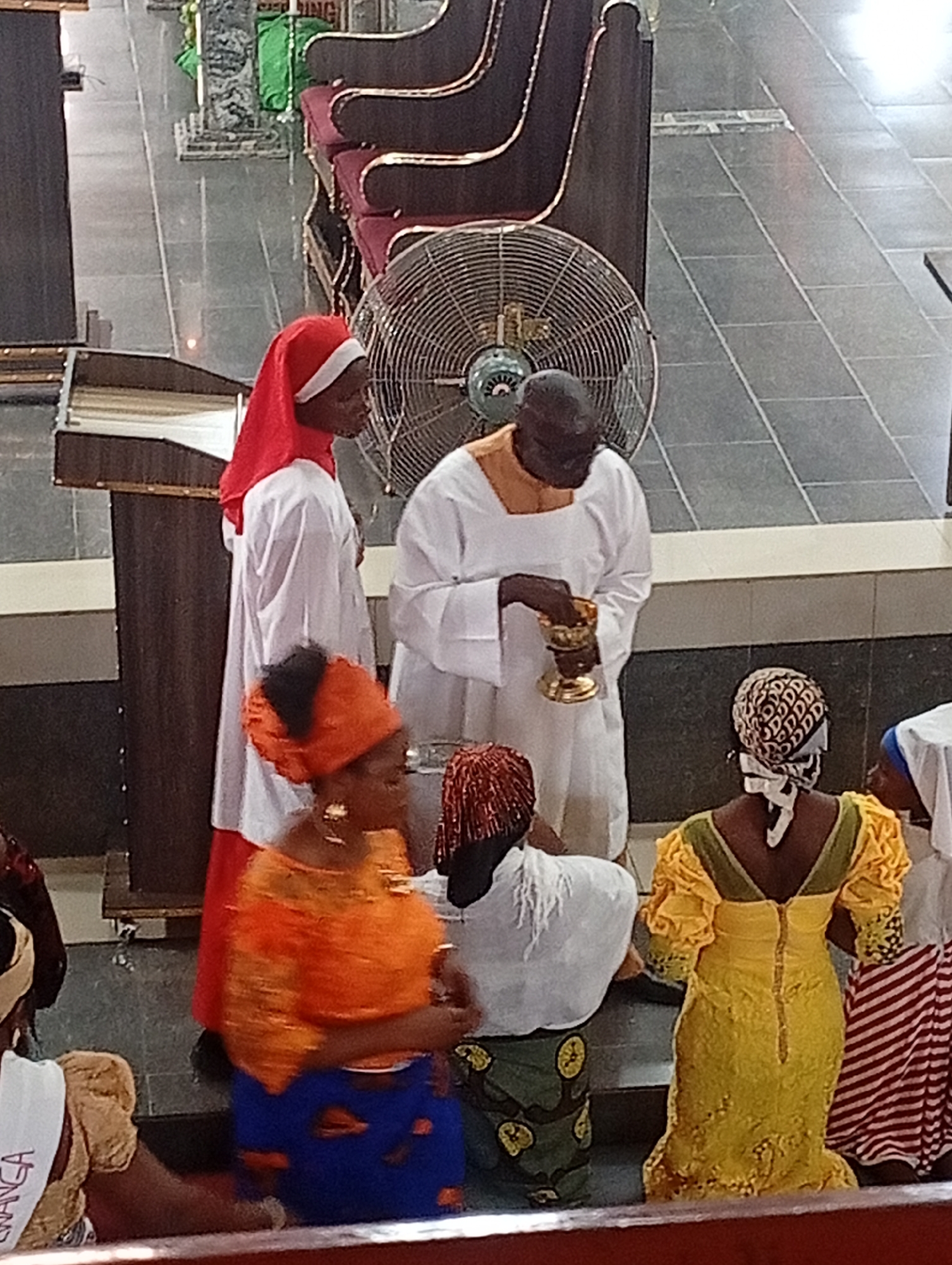 In what appears to be the interior of a religious building in Narayi, Nigeria, a man dressed in white robes holds a golden chalice in his left hand. Standing next to him is another individual wearing a white robe and a red head covering. Several women are kneeling in front of them, dressed in colorful African prints and patterns. A large electric fan is visible in the background. The floor is tiled in a gray pattern. Rows of chairs are seen in the background.