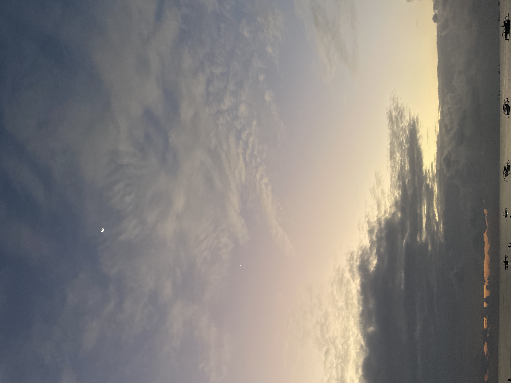The image depicts a serene seascape during what appears to be dusk or dawn. A thin crescent moon is visible in the upper left quadrant of the sky. The sky is a gradient of colors, transitioning from a deep blue at the top to soft hues of pink and yellow closer to the horizon. Clouds are scattered throughout the sky, with some appearing darker and more dense, suggesting thicker cloud cover lower down.
The horizon line is clearly defined, separating the sky from the water. The water itself appears calm, with a few small boats scattered across its surface. These boats are dark silhouettes against the lighter sky and water, and their presence suggests a coastal or marine environment. The overall atmosphere of the image is peaceful and tranquil. The location is identified as Unknown, Unknown.