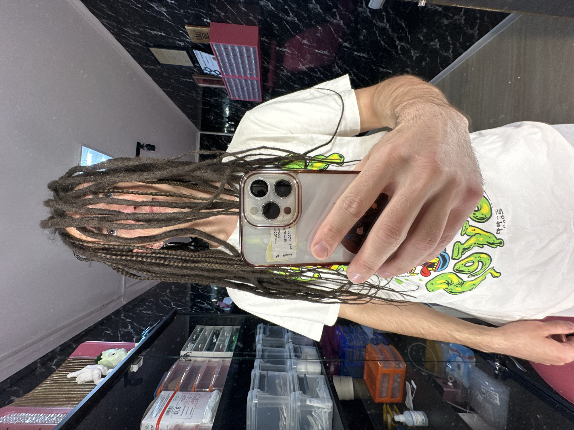 The image is a selfie taken by a person with dreadlocks, partially obscuring their face. They are holding a smartphone with a triple-lens camera in their right hand. The person is wearing a white t-shirt with a green and yellow design. The background suggests a commercial or retail environment. A display case is visible, containing various items, possibly related to optometry or jewelry. The wall in the background has a dark, marble-like pattern. Based on contextual information, the location is in Saratov, Russia, though there are no features in the image to confirm this.