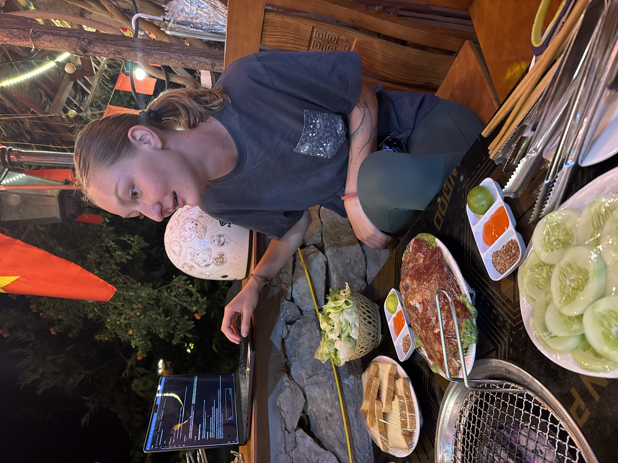 The image shows a woman in a dark gray t-shirt and dark pants sitting at a table in what appears to be a restaurant or outdoor eatery. The table is set with various dishes, including sliced cucumbers, a plate of meat, a basket of greens, and small dishes containing sauces and condiments. A grill is visible on the table, suggesting a barbecue or grilling experience. The woman is looking down at a tablet or screen. In the background, there are trees and vegetation, along with a Vietnamese flag. A white helmet is placed on the ground near the tablet. Based on the presence of the flag and the location name provided, it is reasonable to assume that the setting is Mèo Vạc Commune, Vietnam.