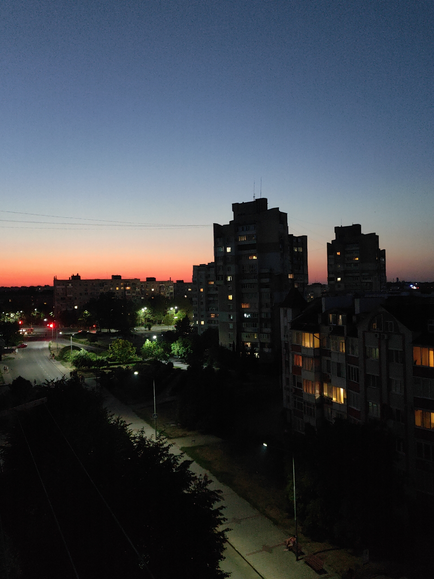 This is an aerial, high-angle, full shot of a residential area in Chornomorsk, Ukraine, at dusk. The sky transitions from a deep blue at the top to a vibrant orange and pink near the horizon. In the foreground, dark trees and shadowed rooftops dominate, with a path illuminated by a streetlamp in the lower right. A winding road with red traffic lights and a few cars and pedestrians is visible in the mid-ground, flanked by more trees and streetlights. Several apartment buildings of varying heights are scattered throughout the scene, with many windows lit up, indicating activity within. The overall impression is that of a quiet urban evening with the last light of day fading.